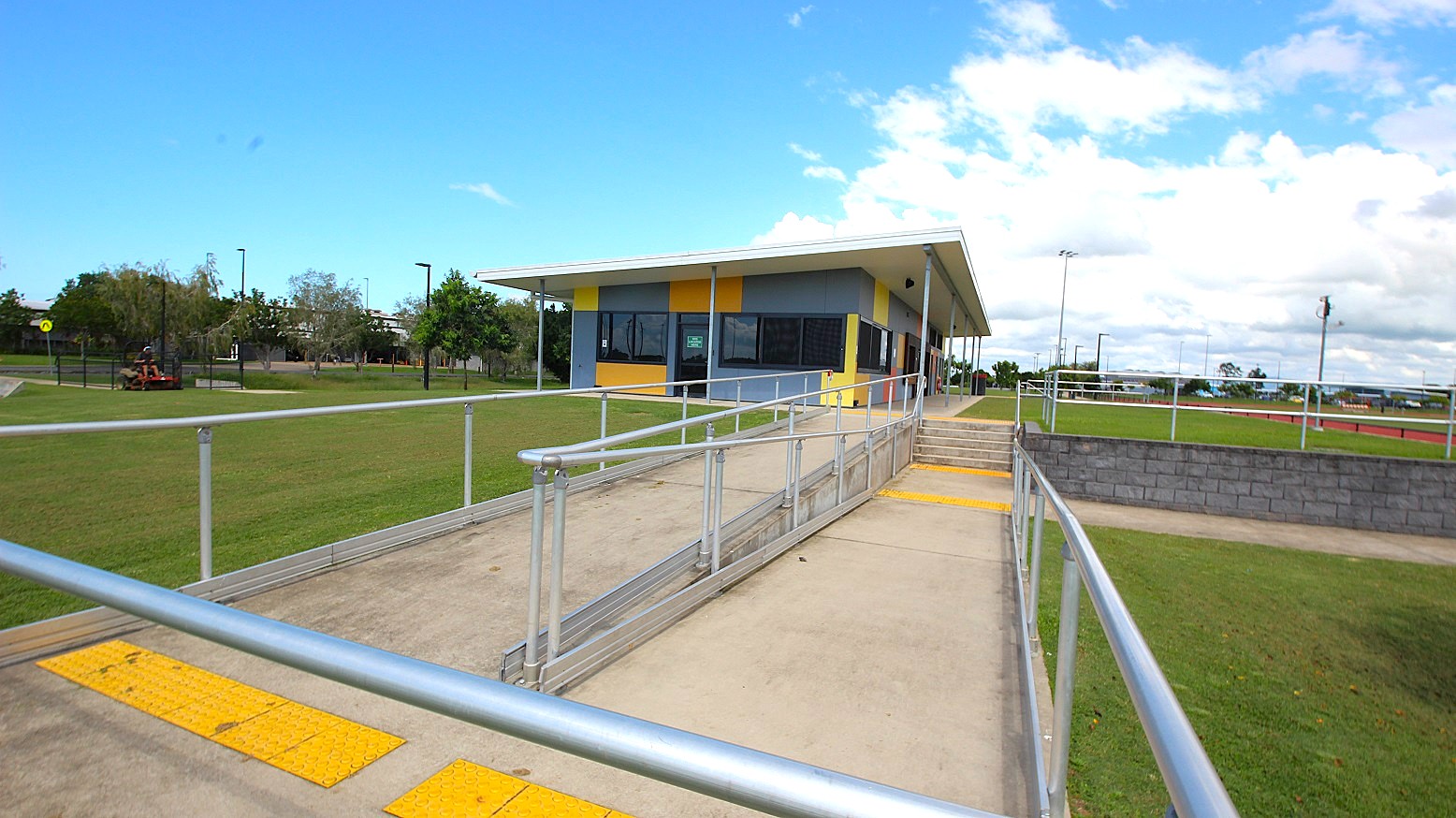 A single-storey building with a sloped roof is in the centre, featuring a colourful facade with blue, yellow, and orange panels. In the foreground, a concrete path with metal railings leads to the building, with a ramp and steps. The surrounding area is grassy, with a few trees and a fence on the left. A low brick wall runs parallel to the path on the right. The sky is partly cloudy. A single-storey building with a sloped roof is in the centre, featuring a colourful facade with blue, yellow, and orange panels. In the foreground, a concrete path with metal railings leads to the building, with a ramp and steps. The surrounding area is grassy, with a few trees and a fence on the left. A low brick wall runs parallel to the path on the right. The sky is partly cloudy.