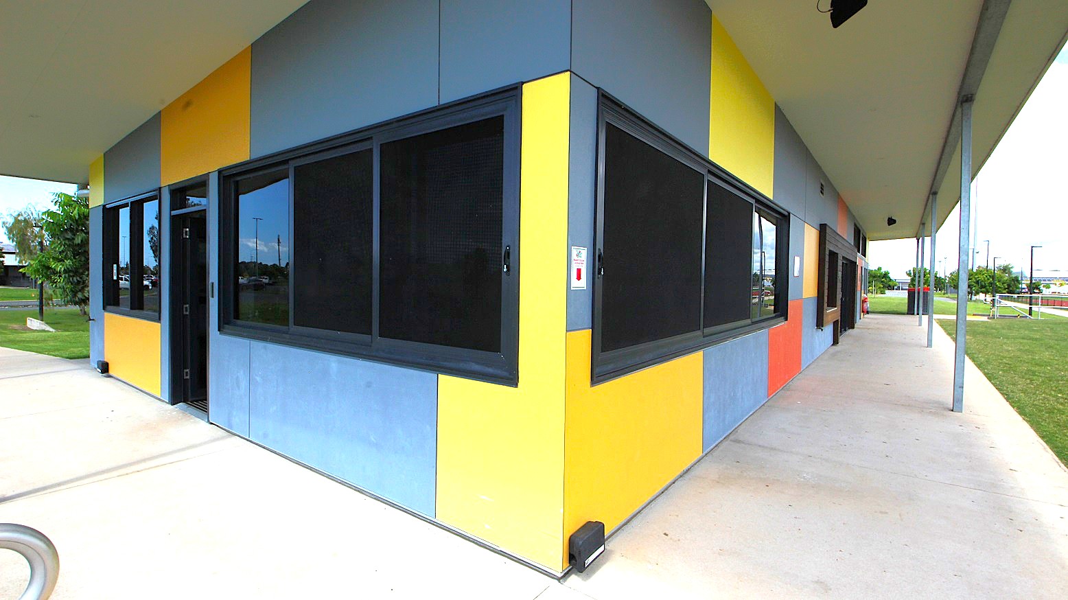 A building with a corner view, featuring a facade of alternating yellow and blue panels. Large, dark-framed windows are set into the walls. The building is situated on a concrete path that extends along its side. The roof is flat and extends outward, supported by metal poles. In the background, there is a grassy area with trees and a fence, and some structures are visible in the distance. A building with a corner view, featuring a facade of alternating yellow and blue panels. Large, dark-framed windows are set into the walls. The building is situated on a concrete path that extends along its side. The roof is flat and extends outward, supported by metal poles. In the background, there is a grassy area with trees and a fence, and some structures are visible in the distance.