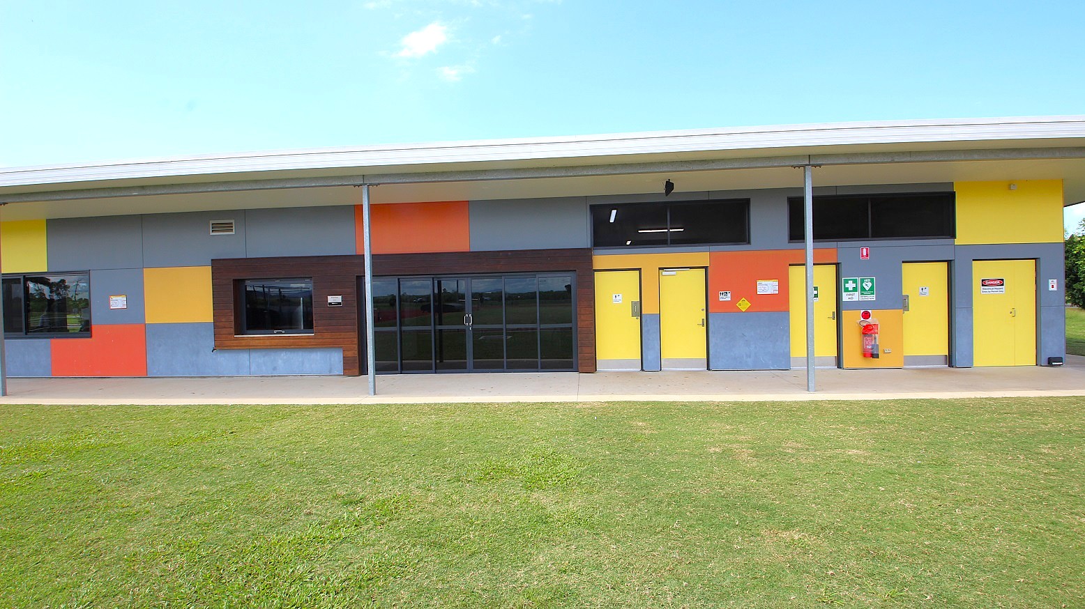 A building with a colourful facade featuring large blocks of yellow, blue, and orange. The structure has several doors and windows, with a prominent set of glass doors in the centre. A fire hydrant is mounted on the wall to the right. The building is supported by metal poles and has a flat roof. In the foreground, there is a well-maintained grassy area. A building with a colourful facade featuring large blocks of yellow, blue, and orange. The structure has several doors and windows, with a prominent set of glass doors in the centre. A fire hydrant is mounted on the wall to the right. The building is supported by metal poles and has a flat roof. In the foreground, there is a well-maintained grassy area.