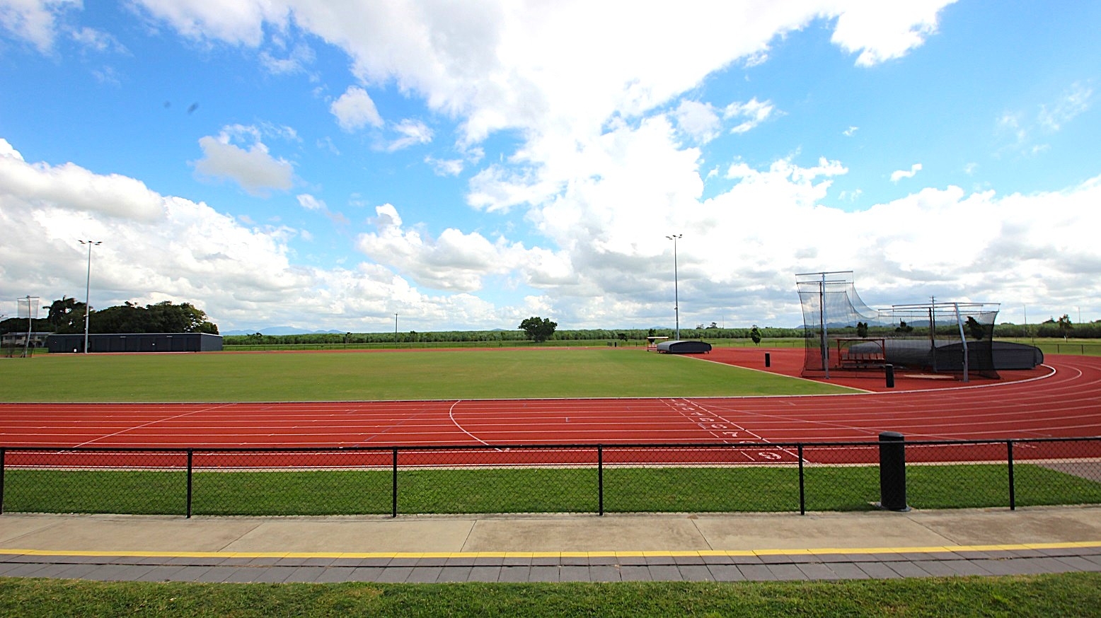 A sports field with a red running track encircling a green grass area. On the right, there is a netted area likely for field events. The track has marked lanes and numbers. In the foreground, a paved path runs parallel to a black metal fence. The background features trees and open fields under a partly cloudy sky. A sports field with a red running track encircling a green grass area. On the right, there is a netted area likely for field events. The track has marked lanes and numbers. In the foreground, a paved path runs parallel to a black metal fence. The background features trees and open fields under a partly cloudy sky.