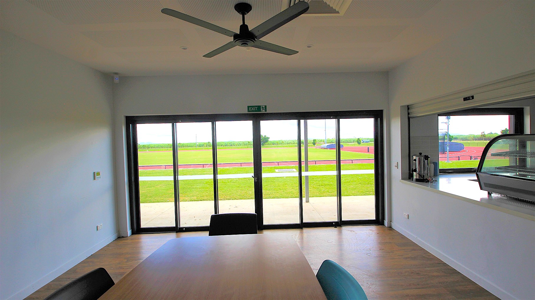 A room with a wooden floor and a ceiling fan. Large glass sliding doors in the centre lead to an outdoor area with a green field and a red running track. On the right, there's a counter with a glass display case and some condiments. A table with chairs is partially visible in the foreground. A room with a wooden floor and a ceiling fan. Large glass sliding doors in the centre lead to an outdoor area with a green field and a red running track. On the right, there's a counter with a glass display case and some condiments. A table with chairs is partially visible in the foreground.