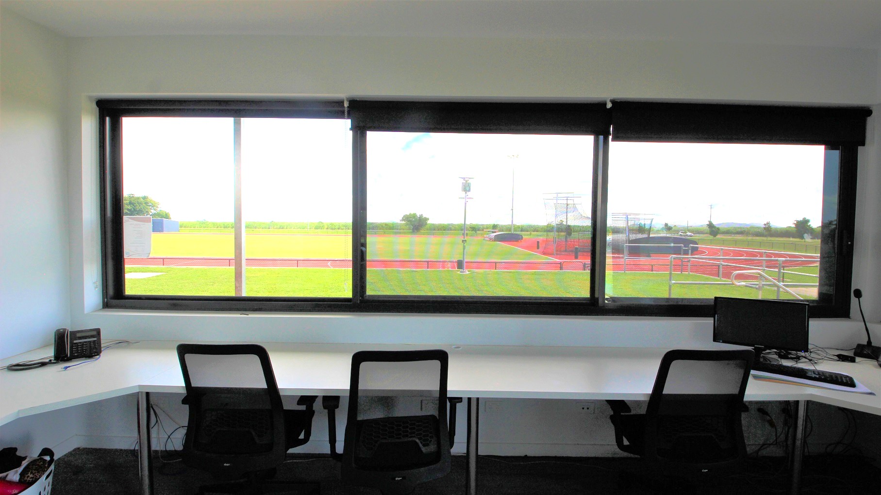 A room with a large window overlooking a sports field with a running track. Inside, there are two black mesh office chairs at a white corner desk. On the desk, a small radio is on the left, and a computer monitor with a keyboard and mouse is on the right. The floor is carpeted in dark grey, and the ceiling is plain white. The view outside shows green grass, a red track, and some equipment in the distance. A room with a large window overlooking a sports field with a running track. Inside, there are two black mesh office chairs at a white corner desk. On the desk, a small radio is on the left, and a computer monitor with a keyboard and mouse is on the right. The floor is carpeted in dark grey, and the ceiling is plain white. The view outside shows green grass, a red track, and some equipment in the distance.