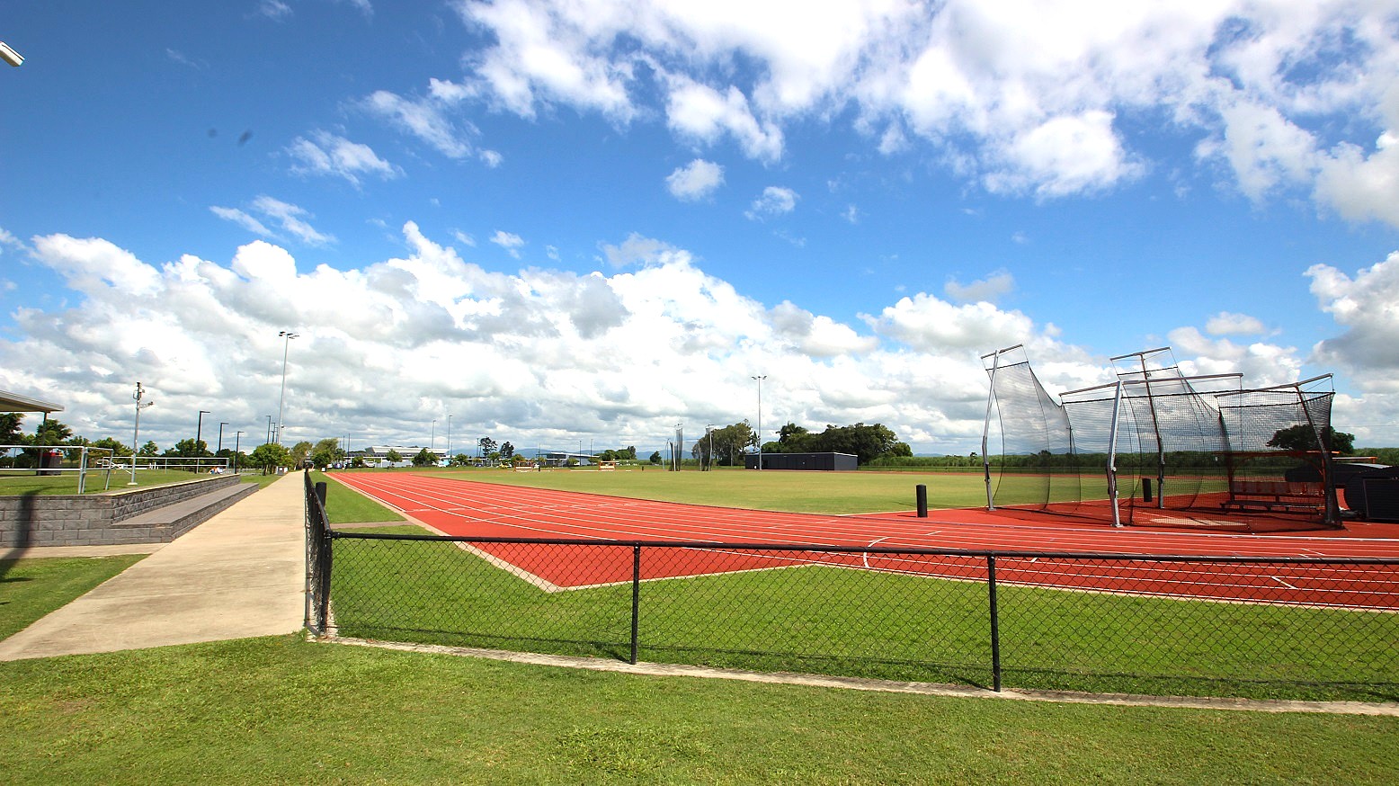 A sports field with a red running track on the right, surrounded by a green grass field. A netted cage structure is visible on the right side of the track. On the left, a concrete pathway runs parallel to the track, with metal railings and a few trees in the distance. The sky is filled with scattered white clouds. A chain-link fence separates the track from the grass in the foreground. A sports field with a red running track on the right, surrounded by a green grass field. A netted cage structure is visible on the right side of the track. On the left, a concrete pathway runs parallel to the track, with metal railings and a few trees in the distance. The sky is filled with scattered white clouds. A chain-link fence separates the track from the grass in the foreground.
