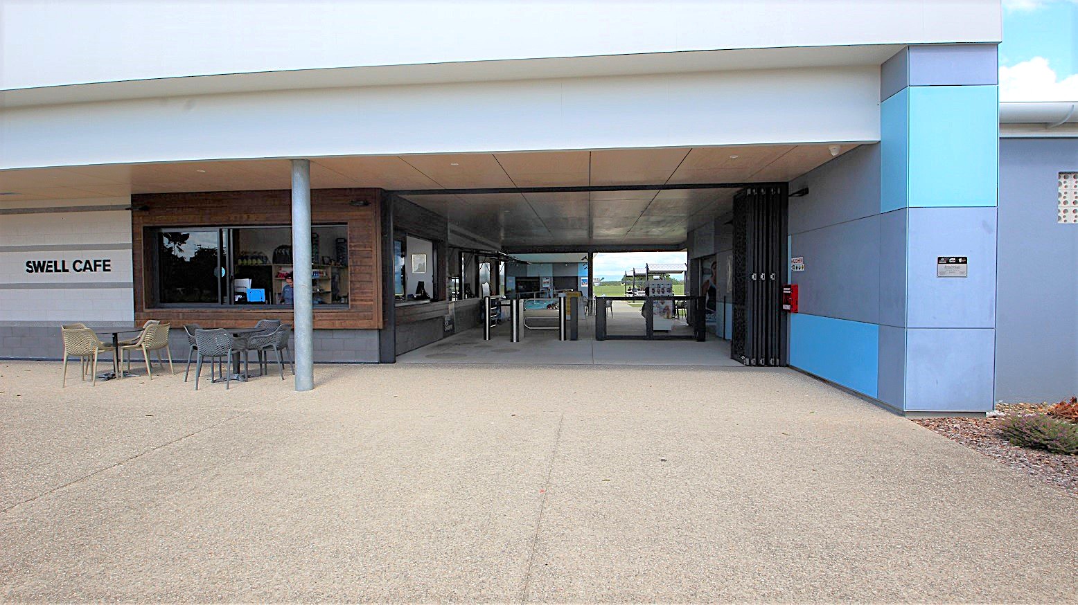Tables and chairs for exterior dining are placed on a paved area in front of the cafe entrance. A wide serving counter with a glass window is visible. The left internal wall has a timber finish. The cafe name sign is attached to the left side. A ticketed entry turnstile and a folding security shutter are positioned to the right of the doorway. A fire extinguisher is mounted on the external wall alongside the entrance. The concrete exterior is painted white, with blue and grey trim accents. Tables and chairs for exterior dining are placed on a paved area in front of the cafe entrance. A wide serving counter with a glass window is visible. The left internal wall has a timber finish. The cafe name sign is attached to the left side. A ticketed entry turnstile and a folding security shutter are positioned to the right of the doorway. A fire extinguisher is mounted on the external wall alongside the entrance. The concrete exterior is painted white, with blue and grey trim accents.