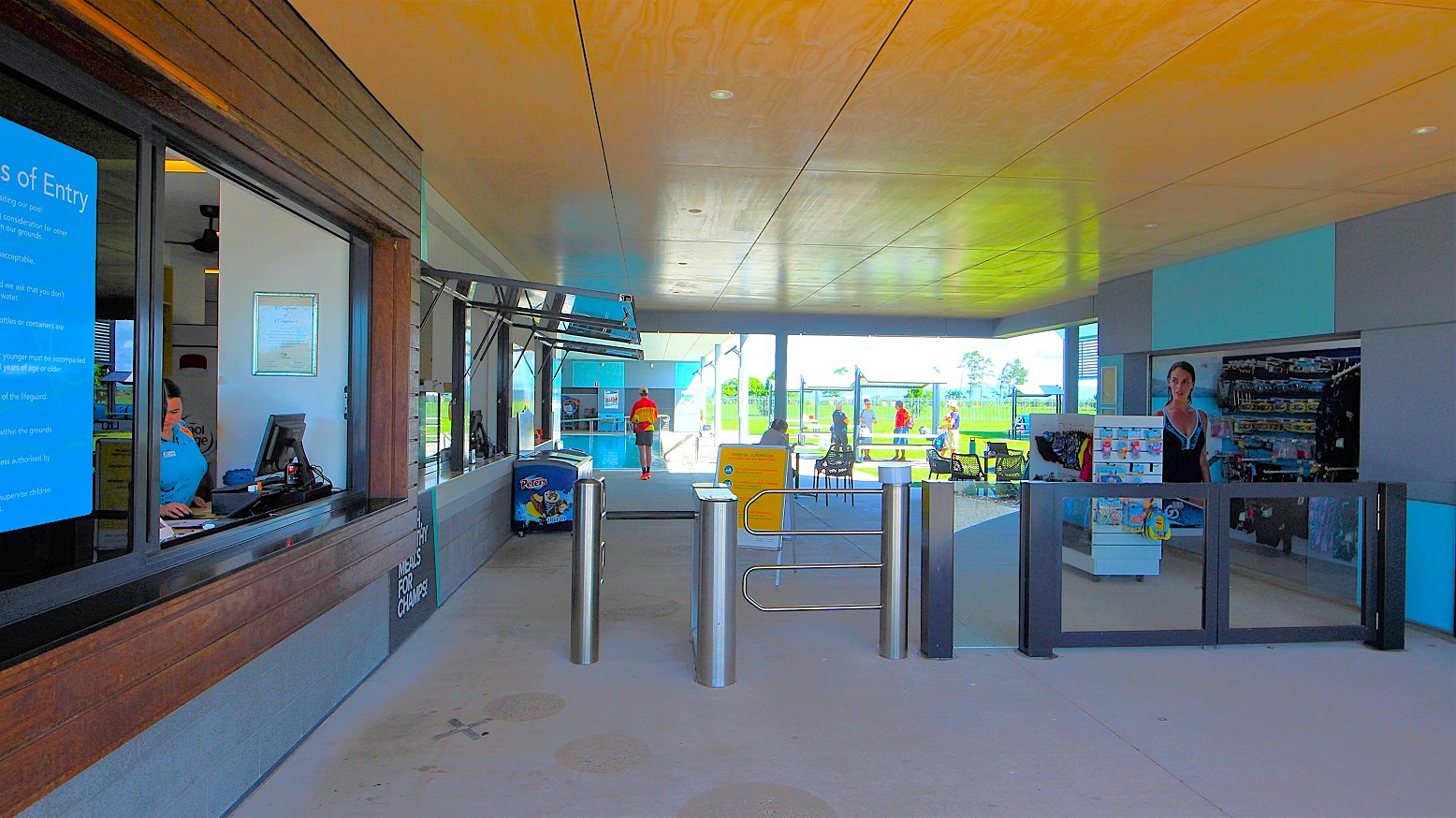 On the left, a reception desk with a wooden front displays a prominent entry condition poster. A staff member stands at a computer. To the right, turnstile gates are visible. Nearby, a small café features an ice cream freezer and a yellow sign. Moveable tables and chairs are arranged. On the right, a retail area showcases swimming goggles and accessories. Further ahead, guests stand under shade structures. On the left, a reception desk with a wooden front displays a prominent entry condition poster. A staff member stands at a computer. To the right, turnstile gates are visible. Nearby, a small café features an ice cream freezer and a yellow sign. Moveable tables and chairs are arranged. On the right, a retail area showcases swimming goggles and accessories. Further ahead, guests stand under shade structures.