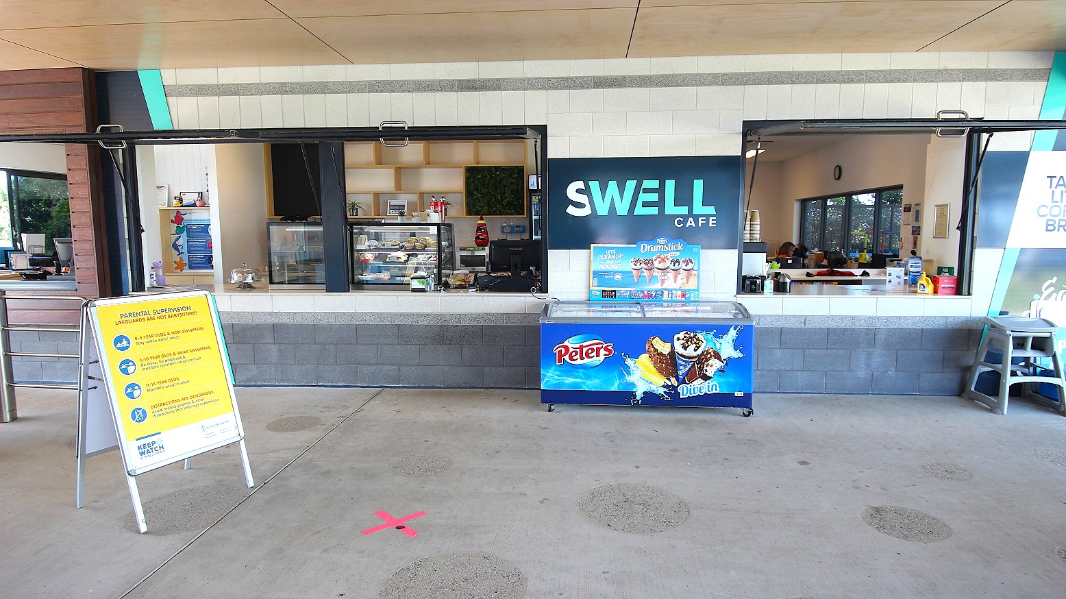 The Swell Cafe displays a grey and white brick facade with a central banner and an ice cream freezer. Inside, food display racks adorn the counter. Wall decorations enhance the ambience. A hot drink dispenser with stacked paper cups is situated on the right. A large banner and a child's high chair are against the wall. A yellow signage is also placed in front.ambi The Swell Cafe displays a grey and white brick facade with a central banner and an ice cream freezer. Inside, food display racks adorn the counter. Wall decorations enhance the ambience. A hot drink dispenser with stacked paper cups is situated on the right. A large banner and a child's high chair are against the wall. A yellow signage is also placed in front.ambi