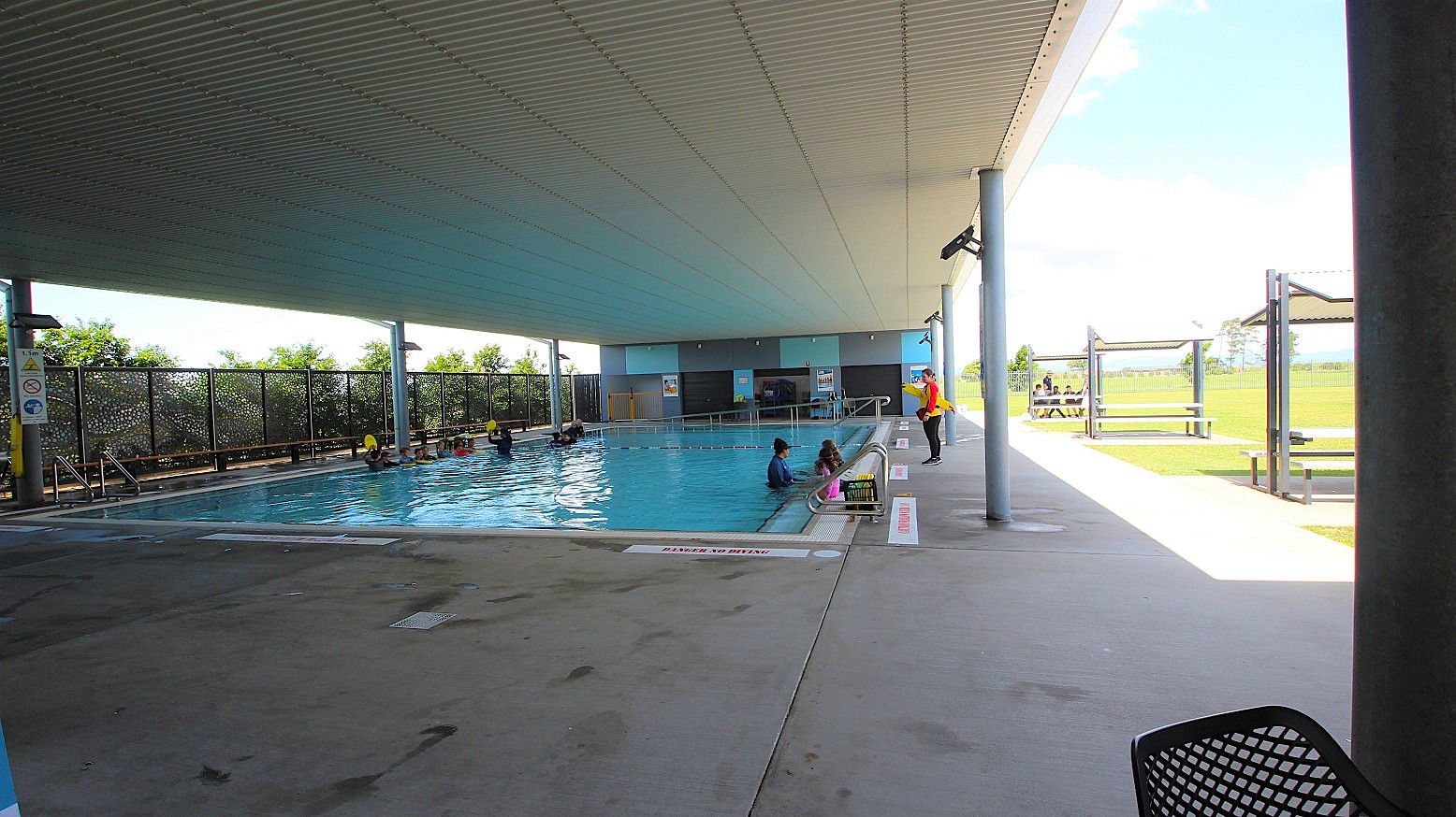 In the centre of an open area lies a pool with a ramp, stairs, and handrails. A metal fence borders the left, accompanied by trees. On the right, shade structures cover picnic tables set on well-maintained green grass. The white corrugated ceiling contrasts with safety banners on the walls. Guests swim while a lifeguard watches over. In the centre of an open area lies a pool with a ramp, stairs, and handrails. A metal fence borders the left, accompanied by trees. On the right, shade structures cover picnic tables set on well-maintained green grass. The white corrugated ceiling contrasts with safety banners on the walls. Guests swim while a lifeguard watches over.
