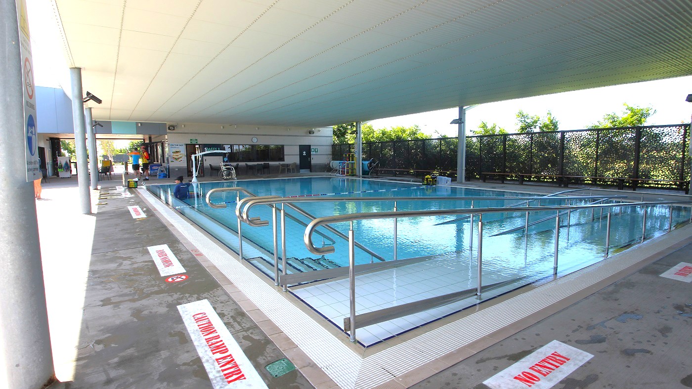 Indoor swimming pool with a ramp leading into the water, surrounded by metal railings. The pool area is covered by a ceiling with a grid pattern. On the left, a walkway with signs on the floor reads "NO ENTRY." In the background, there are people near a seating area and a wall with windows. The right side features a decorative screen with greenery visible through it. The floor is concrete, and the pool is calm and reflective. Indoor swimming pool with a ramp leading into the water, surrounded by metal railings. The pool area is covered by a ceiling with a grid pattern. On the left, a walkway with signs on the floor reads "NO ENTRY." In the background, there are people near a seating area and a wall with windows. The right side features a decorative screen with greenery visible through it. The floor is concrete, and the pool is calm and reflective.