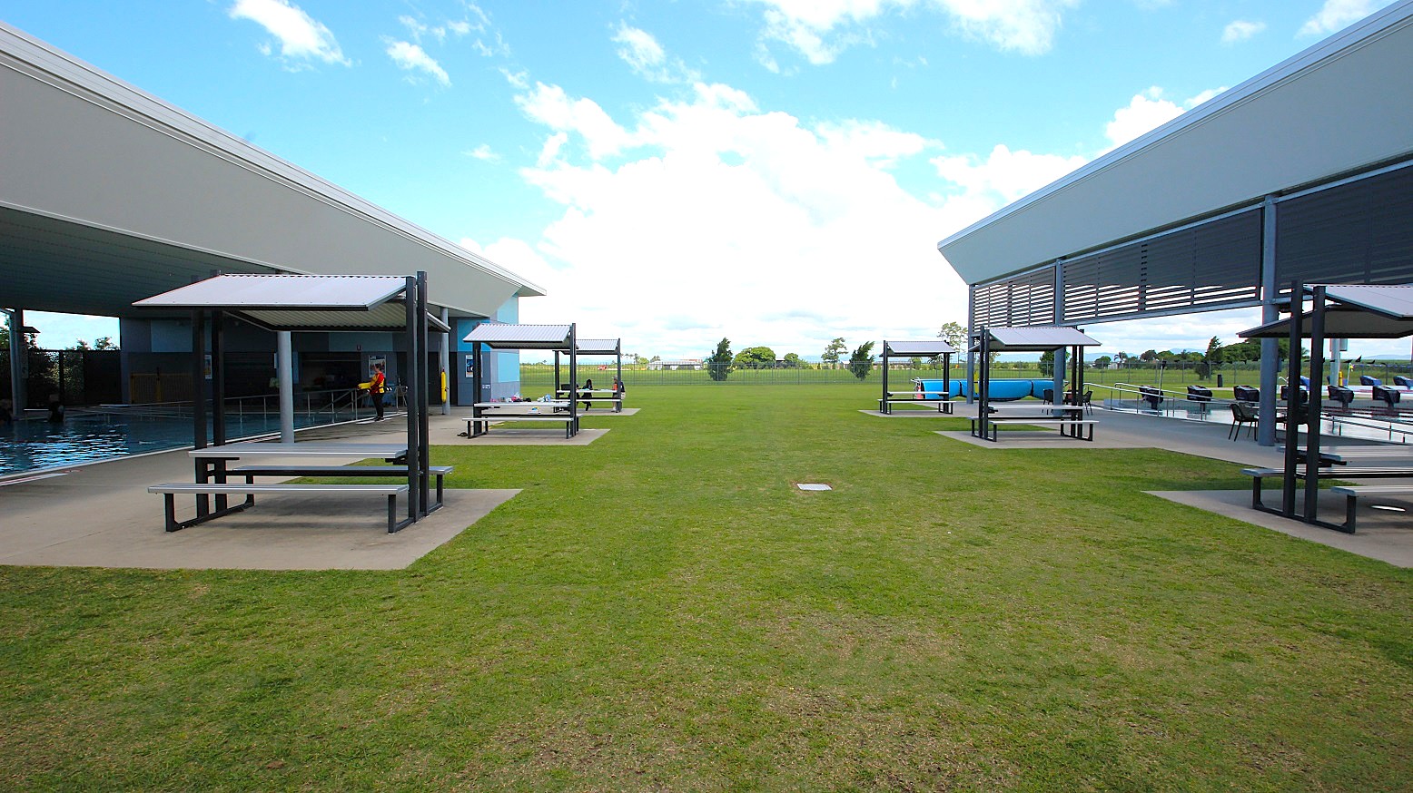 On the left, there is a covered area with a swimming pool and a shaded seating area with benches. On the right, another covered area features similar shaded seating. The middle ground is a grassy open space leading to a distant view of trees and a clear sky. The foreground shows a well-maintained lawn, and the structures have modern, angular designs. On the left, there is a covered area with a swimming pool and a shaded seating area with benches. On the right, another covered area features similar shaded seating. The middle ground is a grassy open space leading to a distant view of trees and a clear sky. The foreground shows a well-maintained lawn, and the structures have modern, angular designs.