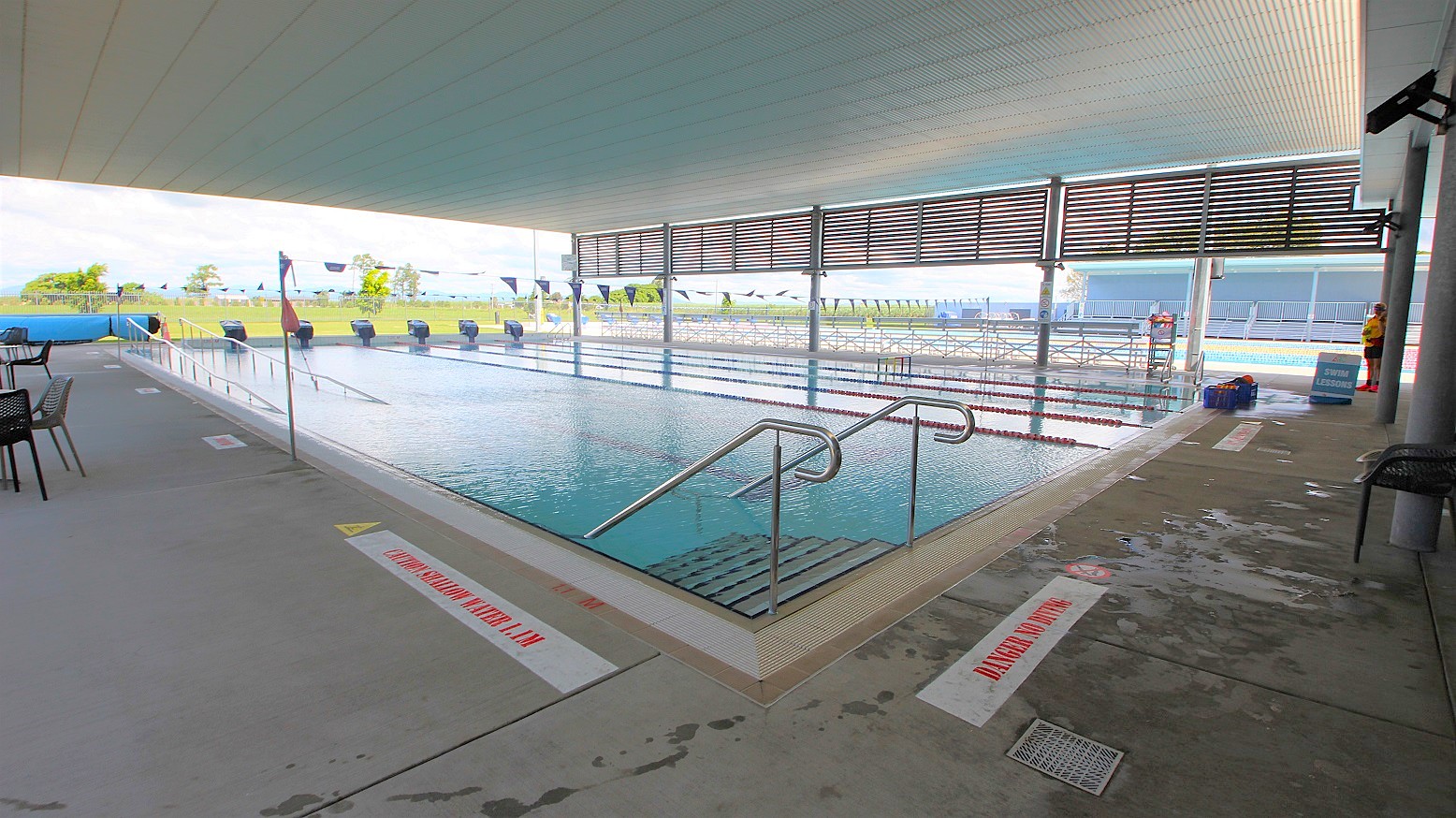 An indoor swimming pool with multiple lanes, bordered by a concrete deck. On the left, there are tables and chairs. The pool has metal handrails leading into the water. The ceiling is covered, and the area is well-lit. In the background, there are large windows with horizontal slats, allowing natural light to enter. Outside, a grassy area and some trees are visible. The floor is wet with visible caution signs. An indoor swimming pool with multiple lanes, bordered by a concrete deck. On the left, there are tables and chairs. The pool has metal handrails leading into the water. The ceiling is covered, and the area is well-lit. In the background, there are large windows with horizontal slats, allowing natural light to enter. Outside, a grassy area and some trees are visible. The floor is wet with visible caution signs.