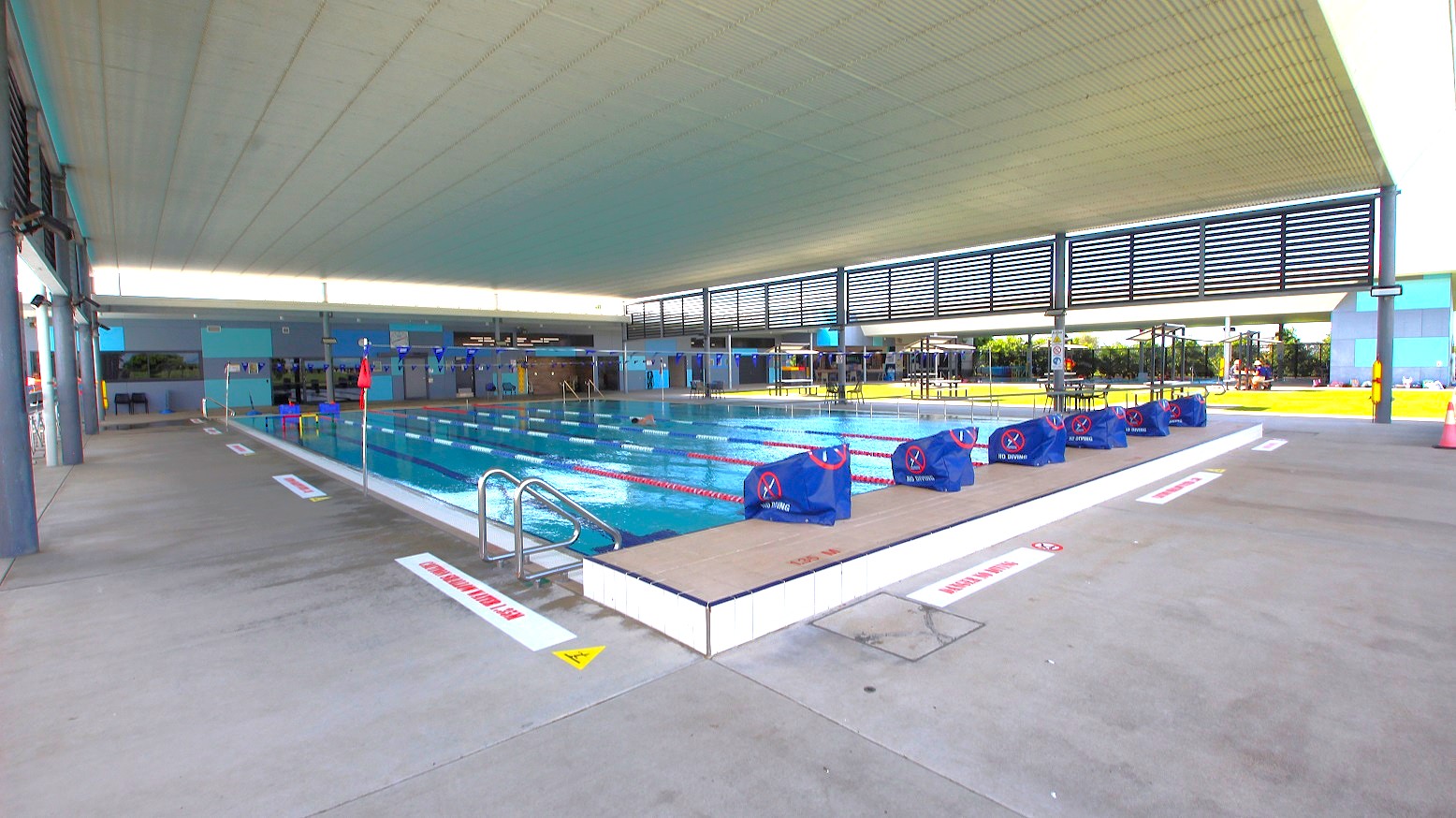 An indoor swimming pool with multiple lanes, separated by red and white lane dividers. Blue starting blocks are positioned at one end. The pool is surrounded by a concrete deck with safety signs. The ceiling is high and white, with slatted panels allowing natural light. On the right, there are tables and chairs on a grassy area visible through large windows. The left side has a walkway with structural columns. An indoor swimming pool with multiple lanes, separated by red and white lane dividers. Blue starting blocks are positioned at one end. The pool is surrounded by a concrete deck with safety signs. The ceiling is high and white, with slatted panels allowing natural light. On the right, there are tables and chairs on a grassy area visible through large windows. The left side has a walkway with structural columns.