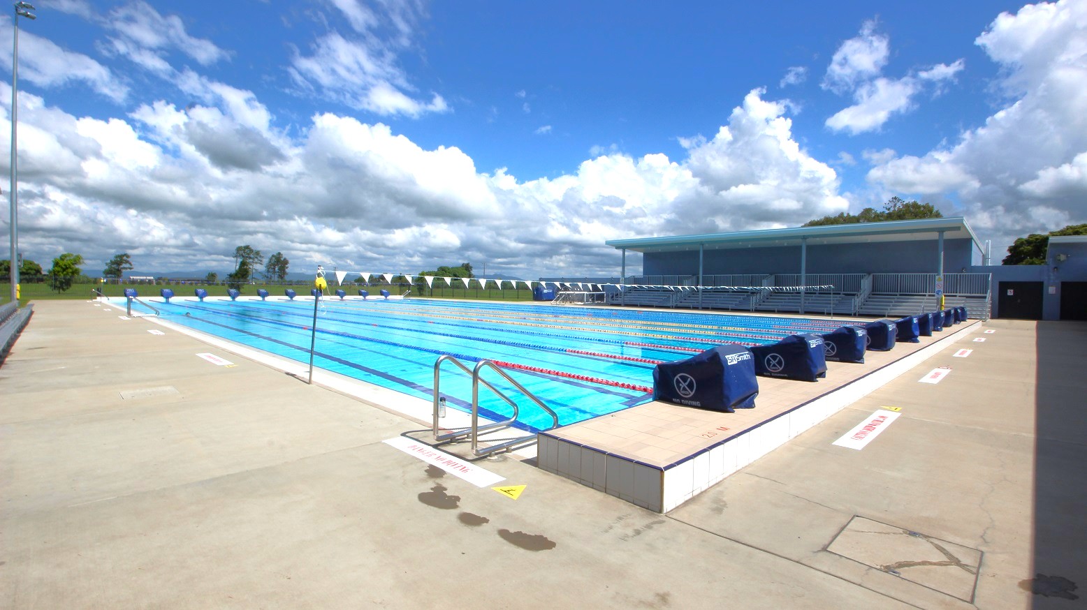 An outdoor swimming pool with multiple lanes, bordered by a concrete deck. On the right, there are covered seating areas with blue covers on the lane dividers. The left side shows an open area with a few trees in the distance. The pool has metal ladders for entry and exit. The sky is filled with scattered clouds. An outdoor swimming pool with multiple lanes, bordered by a concrete deck. On the right, there are covered seating areas with blue covers on the lane dividers. The left side shows an open area with a few trees in the distance. The pool has metal ladders for entry and exit. The sky is filled with scattered clouds.