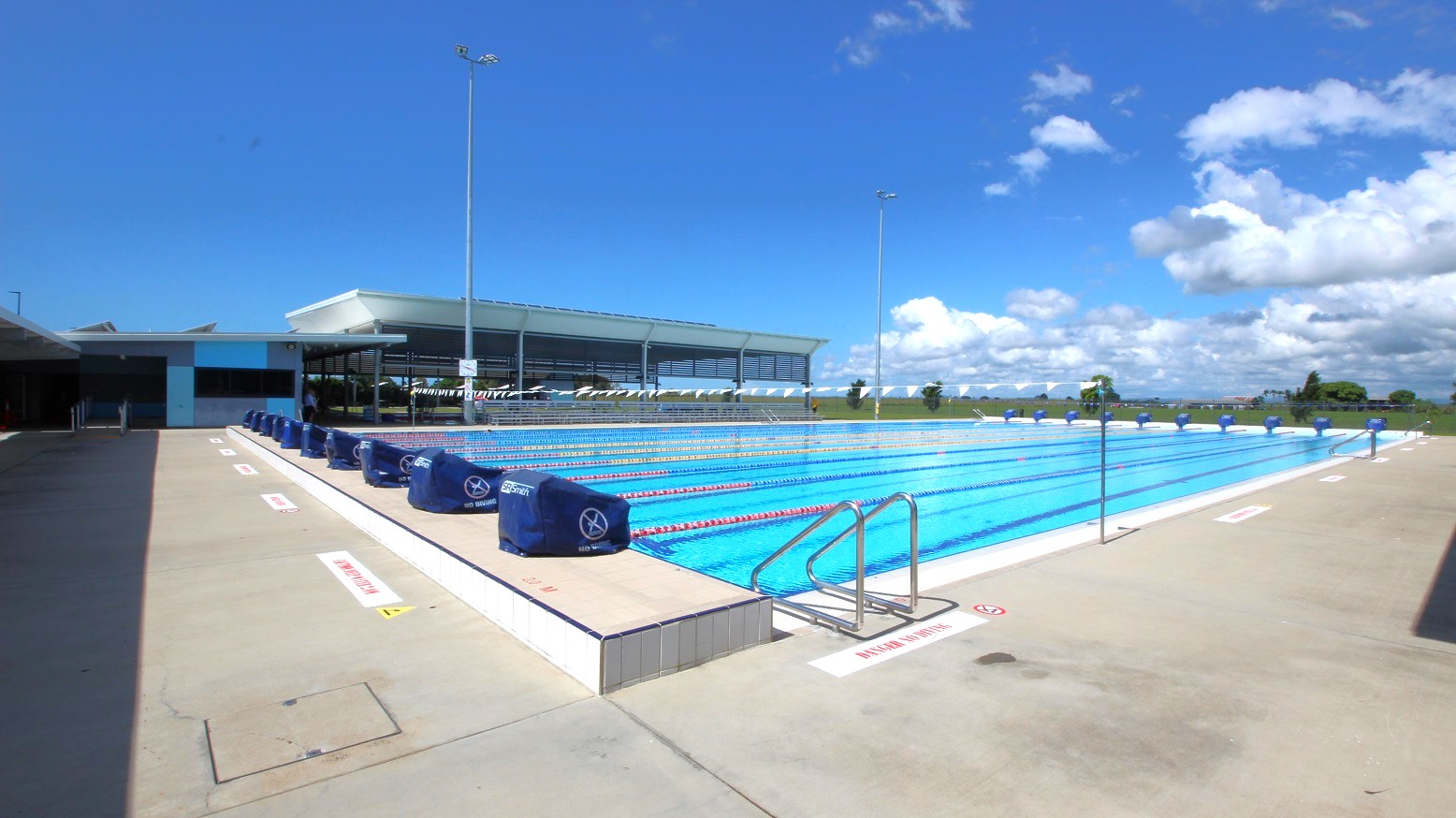 An outdoor swimming pool with multiple lanes, each marked by lane dividers. On the left, there is a shaded walkway leading to a building with a flat roof. The pool deck is concrete, with metal handrails at the pool's edge. In the background, a low building with large windows and a flat roof is visible. The pool is surrounded by open space with some trees and a clear sky above. An outdoor swimming pool with multiple lanes, each marked by lane dividers. On the left, there is a shaded walkway leading to a building with a flat roof. The pool deck is concrete, with metal handrails at the pool's edge. In the background, a low building with large windows and a flat roof is visible. The pool is surrounded by open space with some trees and a clear sky above.