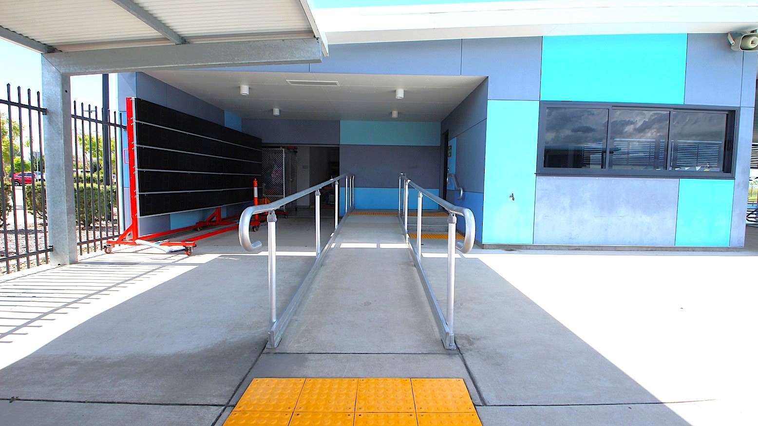 A concrete ramp with metal railings leads into a covered area. On the left, there's a blackboard on a red stand and a black metal fence. On the right, a building with blue and grey panels features a window with closed blinds. The ceiling is flat and grey, with lights installed. The floor has a yellow tactile paving section at the ramp's entrance. A concrete ramp with metal railings leads into a covered area. On the left, there's a blackboard on a red stand and a black metal fence. On the right, a building with blue and grey panels features a window with closed blinds. The ceiling is flat and grey, with lights installed. The floor has a yellow tactile paving section at the ramp's entrance.