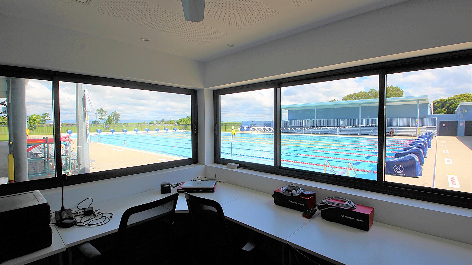 View from inside a room with large windows overlooking an outdoor swimming pool. The pool has multiple lanes with red and blue lane dividers. On the right, there are blue covers on the poolside equipment. The foreground shows a desk with electronic equipment, cables, and a chair. Outside, there is a grassy area and trees in the background. The ceiling is plain, and the floor is not visible. View from inside a room with large windows overlooking an outdoor swimming pool. The pool has multiple lanes with red and blue lane dividers. On the right, there are blue covers on the poolside equipment. The foreground shows a desk with electronic equipment, cables, and a chair. Outside, there is a grassy area and trees in the background. The ceiling is plain, and the floor is not visible.