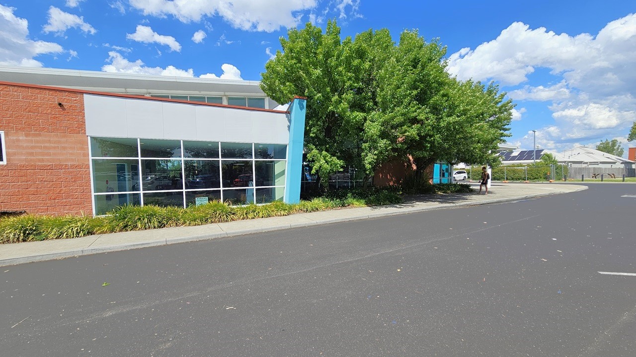 The facility building is a commercial structure featuring red brick, blue-colored concrete, steel, and glass elements. The left side of the building is accompanied by trees and ornamental plants, running parallel to the building and the concrete pathway. The entrance is further ahead, leading to additional commercial buildings equipped with solar panels. A person is standing under the shade of a tree near the curb. The facility building is a commercial structure featuring red brick, blue-colored concrete, steel, and glass elements. The left side of the building is accompanied by trees and ornamental plants, running parallel to the building and the concrete pathway. The entrance is further ahead, leading to additional commercial buildings equipped with solar panels. A person is standing under the shade of a tree near the curb.