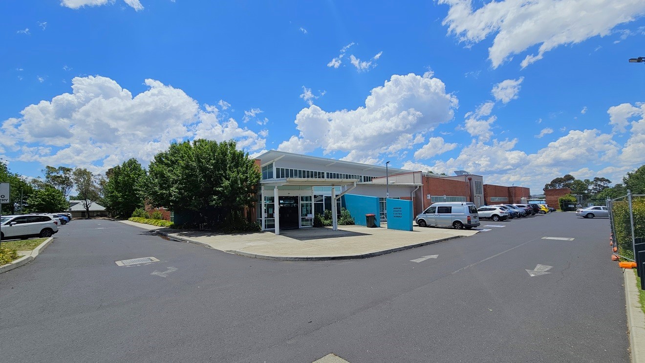 The facility building is a commercial structure featuring red brick, blue-colored concrete, steel, and glass elements. Parking areas, occupied by cars of various colours and models, are situated on both the right and left sides of the building. The surroundings are adorned with trees. The facility building is a commercial structure featuring red brick, blue-colored concrete, steel, and glass elements. Parking areas, occupied by cars of various colours and models, are situated on both the right and left sides of the building. The surroundings are adorned with trees.