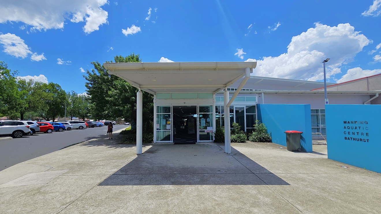 The building entrance features an automatic glass door under a portico. On the left, there is a parking area with cars of various shades and models. To the right is a blue wall displaying the facility's name. A landscape of shrubs is in front of the building, with a rubbish bin and a floodlight positioned on the right near the wall. The building entrance features an automatic glass door under a portico. On the left, there is a parking area with cars of various shades and models. To the right is a blue wall displaying the facility's name. A landscape of shrubs is in front of the building, with a rubbish bin and a floodlight positioned on the right near the wall.