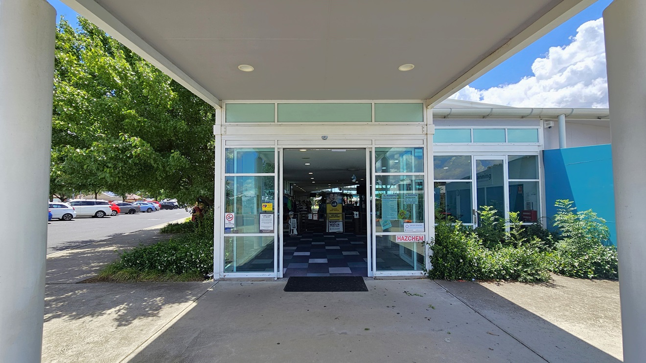 The building entrance has an automatic glass door under a portico, with a non-slip mat on the doorstep. On the left is a parking area with cars of various shades and models. To the right is a blue wall. A landscape of shrubs is in front of the building, with trees and shrubs visible on the left. Inside the building, checked tiles and retail items are visible. The building entrance has an automatic glass door under a portico, with a non-slip mat on the doorstep. On the left is a parking area with cars of various shades and models. To the right is a blue wall. A landscape of shrubs is in front of the building, with trees and shrubs visible on the left. Inside the building, checked tiles and retail items are visible.