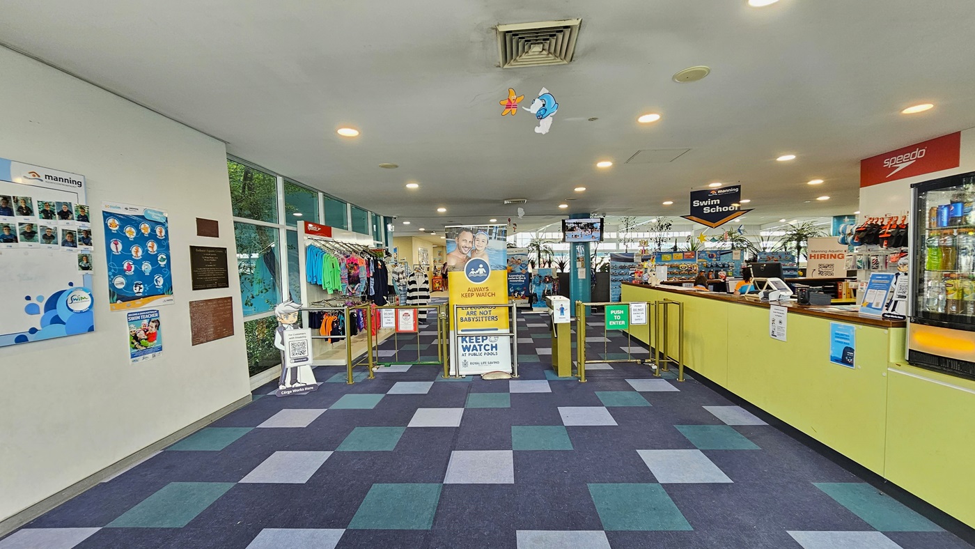 To the right is the reception area with a yellow-front desk, posters, computers, and retail items. A fridge with cold drinks is on its right. A low metal gate serves as a barrier, and a retractable banner stands in the middle. To the left, colorful posters and facility banners adorn the wall. Retail items are displayed after the barrier gates. The ceiling features dolphins and starfish imagery, and the floor has alternating colored carpet tiles. To the right is the reception area with a yellow-front desk, posters, computers, and retail items. A fridge with cold drinks is on its right. A low metal gate serves as a barrier, and a retractable banner stands in the middle. To the left, colorful posters and facility banners adorn the wall. Retail items are displayed after the barrier gates. The ceiling features dolphins and starfish imagery, and the floor has alternating colored carpet tiles.
