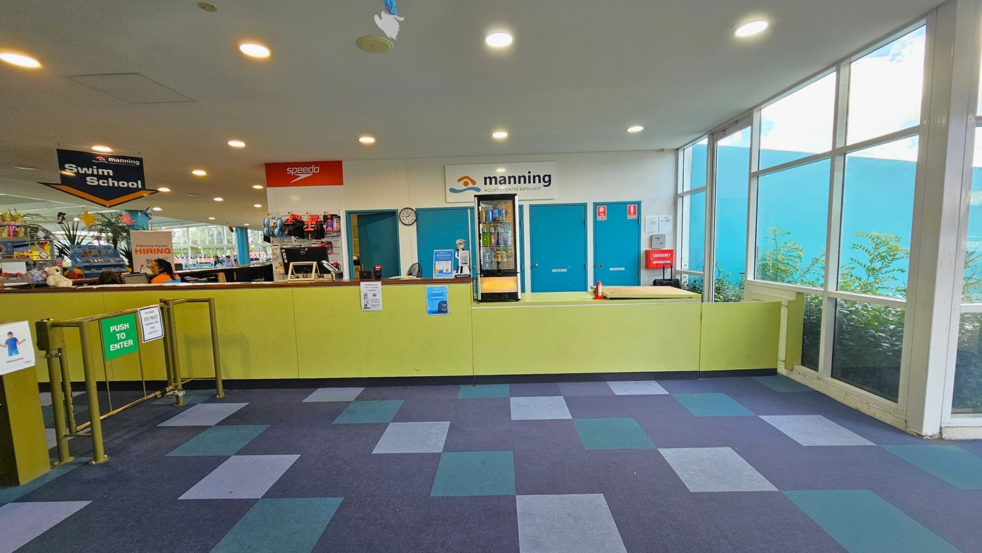 To the right is the reception area with a lower desk, yellow front desk, posters, computers, and floral decorations. Facility banners are also visible. A fridge with cold drinks is behind. A low metal gate serves as a barrier. Retail items are displayed behind the reception desk. The ceiling features dolphins and starfish imagery, and the floor has alternating colored carpet tiles. To the right is the reception area with a lower desk, yellow front desk, posters, computers, and floral decorations. Facility banners are also visible. A fridge with cold drinks is behind. A low metal gate serves as a barrier. Retail items are displayed behind the reception desk. The ceiling features dolphins and starfish imagery, and the floor has alternating colored carpet tiles.