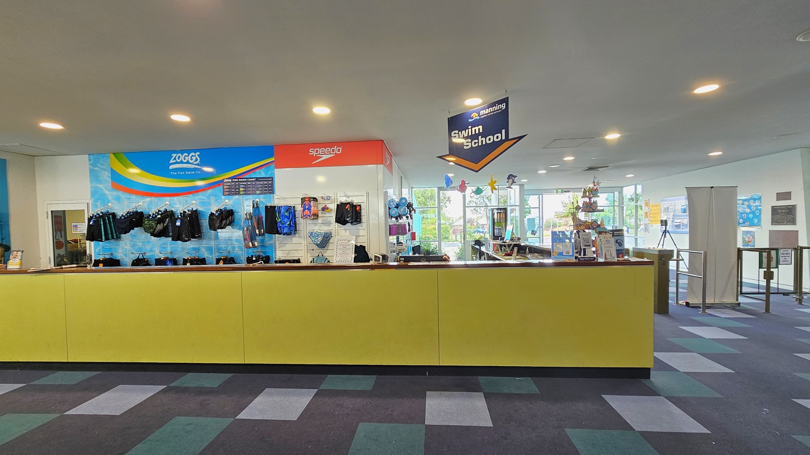 Retail items, including goggles and swimming accessories, are displayed behind the yellow reception desk. To the right is a low gate barrier and a retractable poster. The right wall is adorned with colorful posters and facility banners. The ceiling features all white with LED lights and sea creature imagery, and facility signage is suspended from the ceiling. The floor has alternating colored carpet tiles. Retail items, including goggles and swimming accessories, are displayed behind the yellow reception desk. To the right is a low gate barrier and a retractable poster. The right wall is adorned with colorful posters and facility banners. The ceiling features all white with LED lights and sea creature imagery, and facility signage is suspended from the ceiling. The floor has alternating colored carpet tiles.