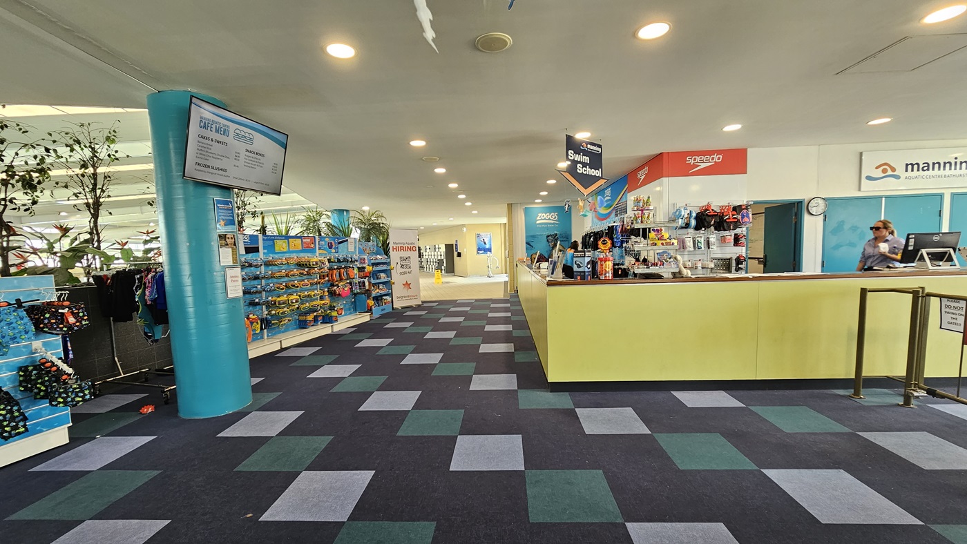 Retail items, such as goggles and swimming accessories, are exhibited behind the yellow reception desk. To the right, there is a low gate barrier and a retractable poster. On the left, tall planter boxes with indoor plants and a long display rack filled with goggles and swimwear are featured. The ceiling is all white with LED lights and facility signage is suspended from the ceiling. A flat-screen TV is mounted to the pillar on the left. The floor is covered with alternating colored carpet tiles Retail items, such as goggles and swimming accessories, are exhibited behind the yellow reception desk. To the right, there is a low gate barrier and a retractable poster. On the left, tall planter boxes with indoor plants and a long display rack filled with goggles and swimwear are featured. The ceiling is all white with LED lights and facility signage is suspended from the ceiling. A flat-screen TV is mounted to the pillar on the left. The floor is covered with alternating colored carpet tiles