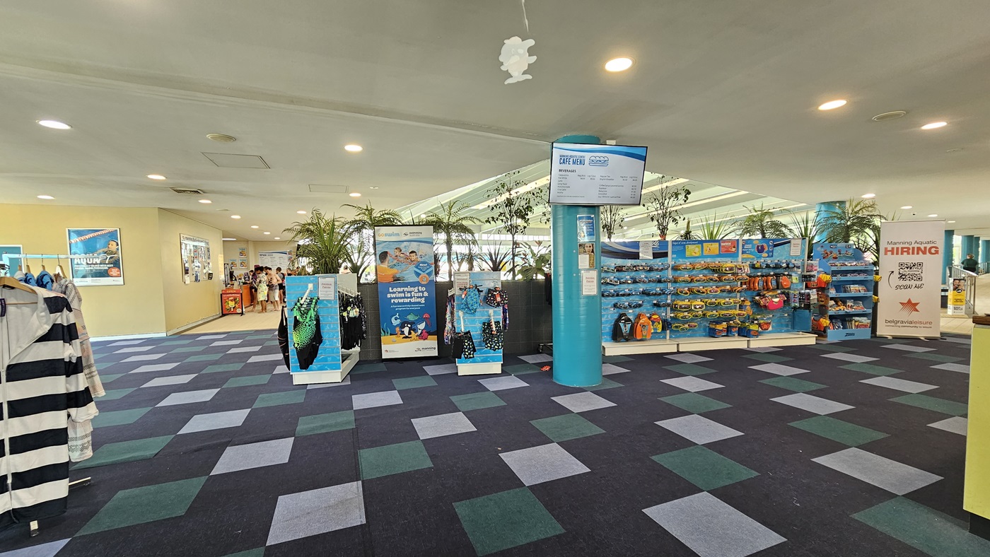 In the middle, tall planter boxes with indoor plants, a display rack filled with goggles, and a swimwear stand. Clothing for sale hangs on the left, and retractable banners are near the planter box. The ceiling is white with LED lights, and facility signage hangs. A flat-screen TV, posters, and people in a corridor on the left are visible. The floor has alternating colored carpet tiles. In the middle, tall planter boxes with indoor plants, a display rack filled with goggles, and a swimwear stand. Clothing for sale hangs on the left, and retractable banners are near the planter box. The ceiling is white with LED lights, and facility signage hangs. A flat-screen TV, posters, and people in a corridor on the left are visible. The floor has alternating colored carpet tiles.