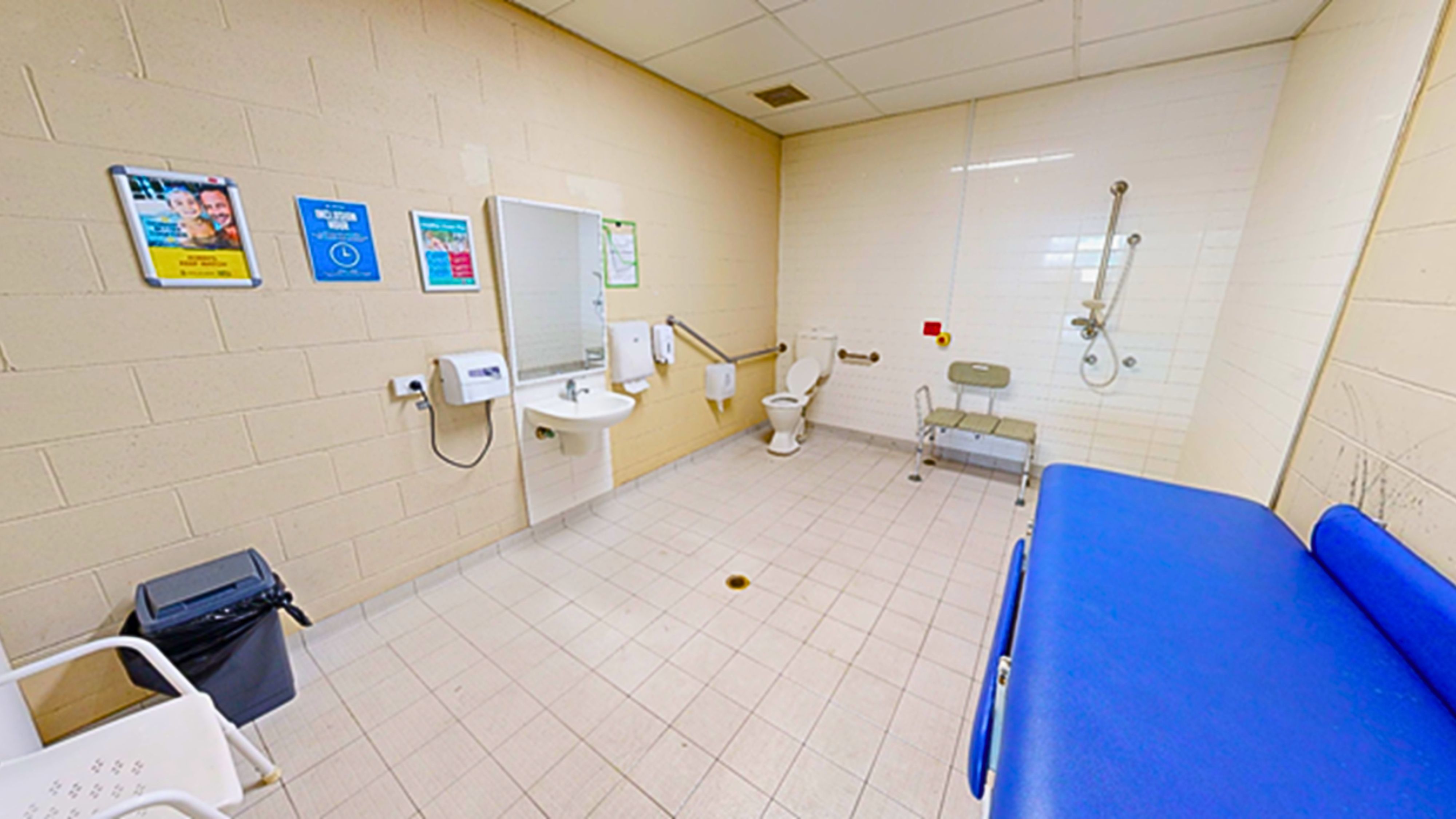 Inside the accessible bathroom, the left corner features a toilet bowl with handrails, a tissue dispenser, and a nearby shower with a bench chair. On the left wall, there's a washbasin, hand dryer, soap and tissue dispenser, and a mirror. Posters are mounted on the wall. A white plastic chair and two rubbish bins are on the left, while a foldable change table is on the right. The floor, ceiling, and wall are covered in white tiles. Inside the accessible bathroom, the left corner features a toilet bowl with handrails, a tissue dispenser, and a nearby shower with a bench chair. On the left wall, there's a washbasin, hand dryer, soap and tissue dispenser, and a mirror. Posters are mounted on the wall. A white plastic chair and two rubbish bins are on the left, while a foldable change table is on the right. The floor, ceiling, and wall are covered in white tiles.