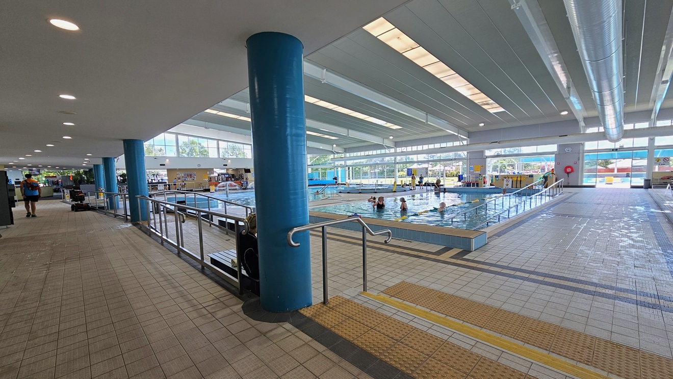 Access to the pool is via a ramp with handrails on the left, and on the right, there's a stepped entry with handrails and truncated domes. The Kids Leisure Pool and training Pool are on the far left, equipped with a ramp and handrail. To the right is the 25m lap pool with ladder access. A pool deck separates the two pools. The floor has matte grey tiles, and the ceiling features strips of transparent roofing, allowing natural sunlight and large aircon vents. Access to the pool is via a ramp with handrails on the left, and on the right, there's a stepped entry with handrails and truncated domes. The Kids Leisure Pool and training Pool are on the far left, equipped with a ramp and handrail. To the right is the 25m lap pool with ladder access. A pool deck separates the two pools. The floor has matte grey tiles, and the ceiling features strips of transparent roofing, allowing natural sunlight and large aircon vents.