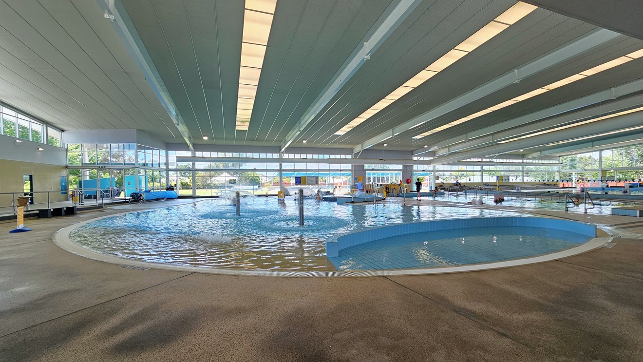 In the middle is the Leisure Pool with a lazy river ride, umbrella water features, and a walled toddler area. The pool is surrounded by an overflow grid. On the left, there is bench seating and a free-standing banner on the pool deck. To the right is the Program Pool equipped with handrails. The area is surrounded by glass, and the ceiling features strips of transparent roofing that allow natural sunlight. In the middle is the Leisure Pool with a lazy river ride, umbrella water features, and a walled toddler area. The pool is surrounded by an overflow grid. On the left, there is bench seating and a free-standing banner on the pool deck. To the right is the Program Pool equipped with handrails. The area is surrounded by glass, and the ceiling features strips of transparent roofing that allow natural sunlight.