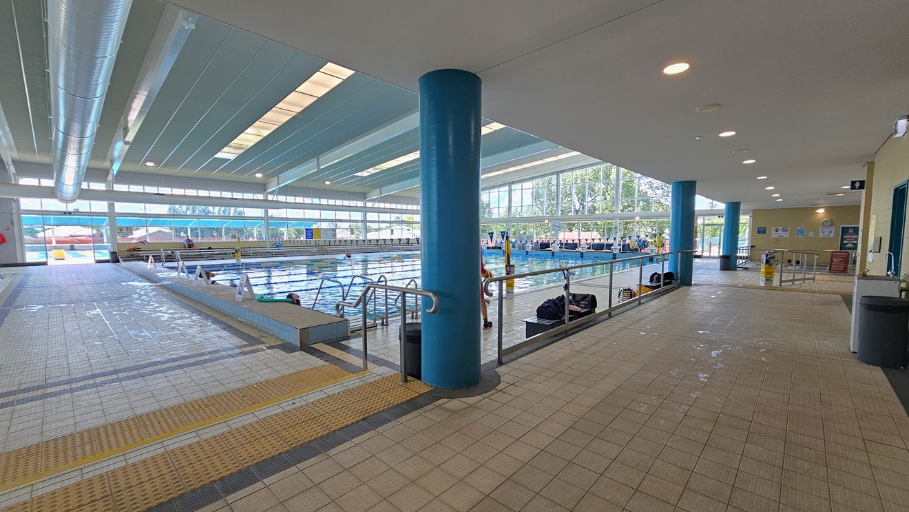 Pool access is via a left ramp with handrails and a right stepped entry with handrails and truncated domes. The 25m lap pool with ladder and handrail access is on the right. Freestanding banners adorn the pool deck. The floor features matte grey tiles, and the ceiling has strips of transparent roofing for natural sunlight, complemented by large aircon vents. The left corridor leads to the bathroom, where a drinking fountain, rubbish bins, and bench chairs are visible. Pool access is via a left ramp with handrails and a right stepped entry with handrails and truncated domes. The 25m lap pool with ladder and handrail access is on the right. Freestanding banners adorn the pool deck. The floor features matte grey tiles, and the ceiling has strips of transparent roofing for natural sunlight, complemented by large aircon vents. The left corridor leads to the bathroom, where a drinking fountain, rubbish bins, and bench chairs are visible.