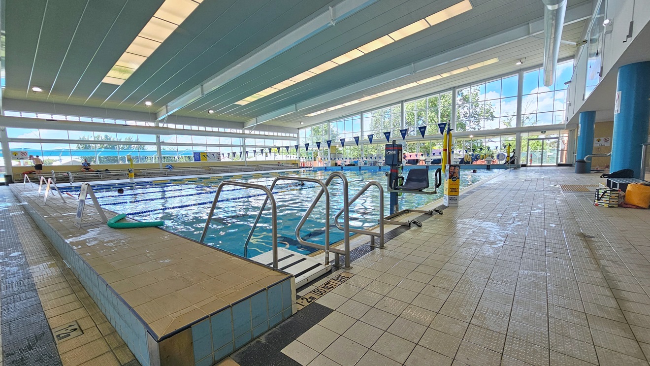 The 25m lap pool features a ladder and handrails, accompanied by an aquatic wheelchair and hoist in the corner. Free Standing banners and a green noodle hose decorate the left pool deck. Matte grey tiles cover the floor, and strips of transparent roofing in the ceiling provide natural sunlight, along with large aircon vents. Tiered seating is available on the far left, and bench chairs are positioned on the right. The 25m lap pool features a ladder and handrails, accompanied by an aquatic wheelchair and hoist in the corner. Free Standing banners and a green noodle hose decorate the left pool deck. Matte grey tiles cover the floor, and strips of transparent roofing in the ceiling provide natural sunlight, along with large aircon vents. Tiered seating is available on the far left, and bench chairs are positioned on the right.