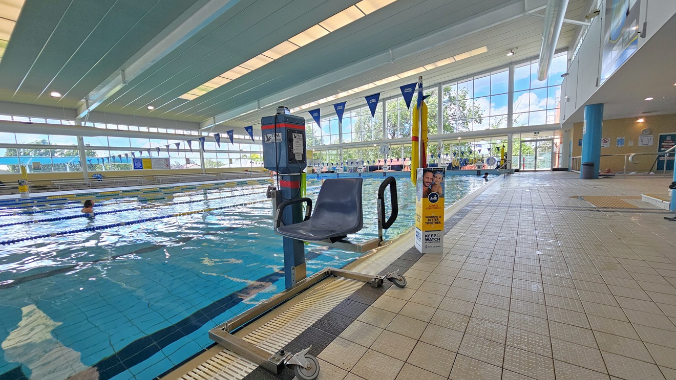 The 25m lap pool is equipped with a ladder and handrails, and the pool deck features an aquatic wheelchair and hoist. Floating ropes and blue bunting banners run across the pool. The high ceiling incorporates strips of transparent roofing, allowing natural sunlight, and large aircon vents. Tiered seating is available on the far left, and the floor is tiled in grey. A glass wall surrounds the area. The 25m lap pool is equipped with a ladder and handrails, and the pool deck features an aquatic wheelchair and hoist. Floating ropes and blue bunting banners run across the pool. The high ceiling incorporates strips of transparent roofing, allowing natural sunlight, and large aircon vents. Tiered seating is available on the far left, and the floor is tiled in grey. A glass wall surrounds the area.