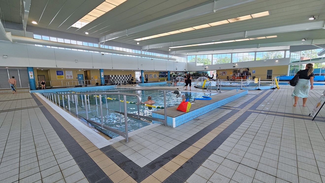 The Program Pool features a ramp with handrails for access, with floating ropes running across the pool. Belongings are visible on the raised pool deck. The Leisure Pool, located to the far right, is equipped with umbrella showers and water features, surrounded by rope bollards. Change places are situated on the opposite side of the pool. The floor has grey tiles, and guests are present in the pool. The surrounding upper wall is made of glass, providing a view of the outside. The Program Pool features a ramp with handrails for access, with floating ropes running across the pool. Belongings are visible on the raised pool deck. The Leisure Pool, located to the far right, is equipped with umbrella showers and water features, surrounded by rope bollards. Change places are situated on the opposite side of the pool. The floor has grey tiles, and guests are present in the pool. The surrounding upper wall is made of glass, providing a view of the outside.