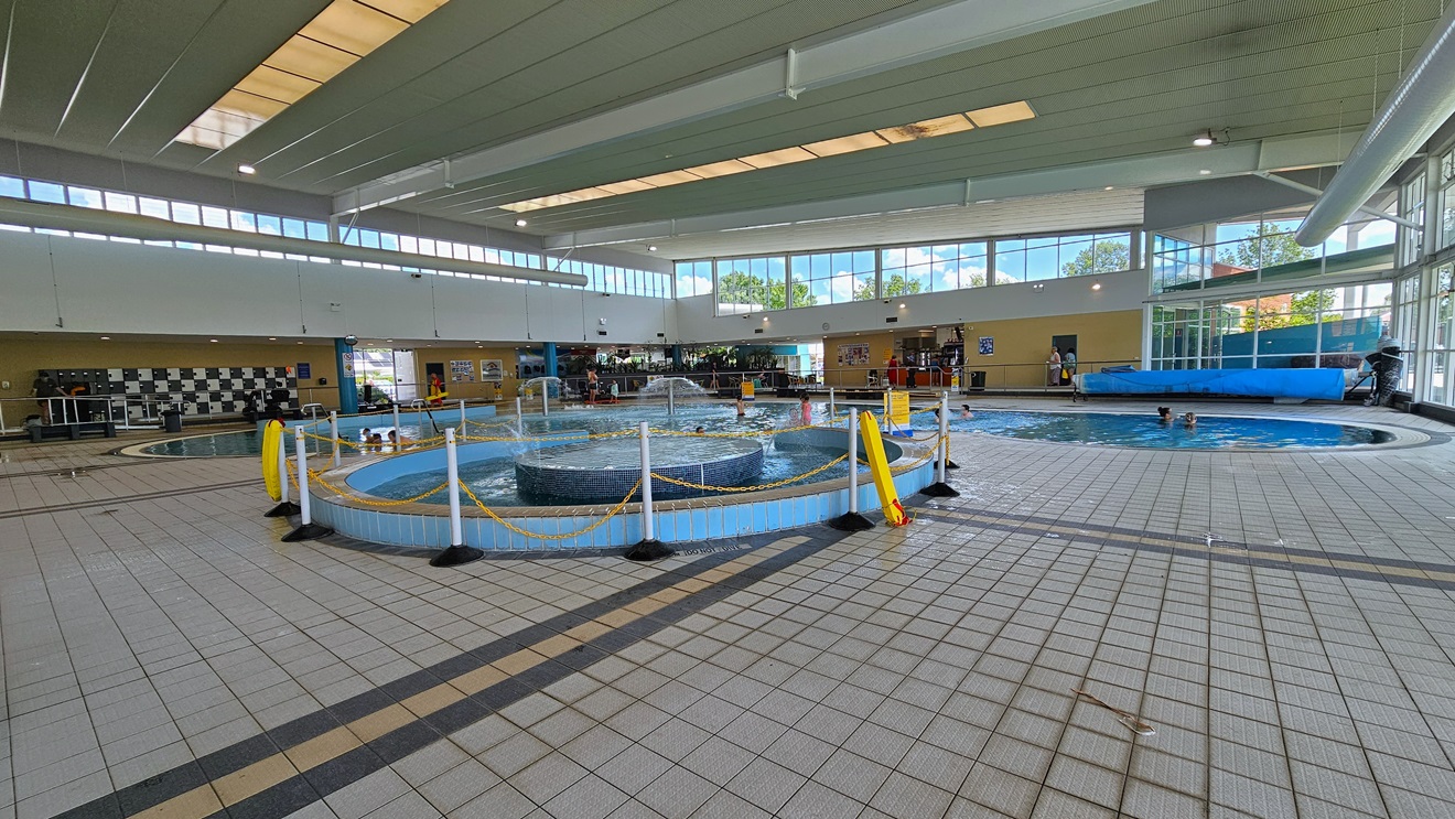 The Leisure Pool includes a lazy river ride with umbrella water features and a walled toddler area. A section of the pool has a rope bollard barricade. To the far left is the locker area. The floor features grey tiles, and guests are present in the pool. The surrounding upper wall is made of glass, providing a view of the outside. The high ceiling incorporates strips of transparent roofing, allowing natural sunlight, and large aircon vents. The Leisure Pool includes a lazy river ride with umbrella water features and a walled toddler area. A section of the pool has a rope bollard barricade. To the far left is the locker area. The floor features grey tiles, and guests are present in the pool. The surrounding upper wall is made of glass, providing a view of the outside. The high ceiling incorporates strips of transparent roofing, allowing natural sunlight, and large aircon vents.