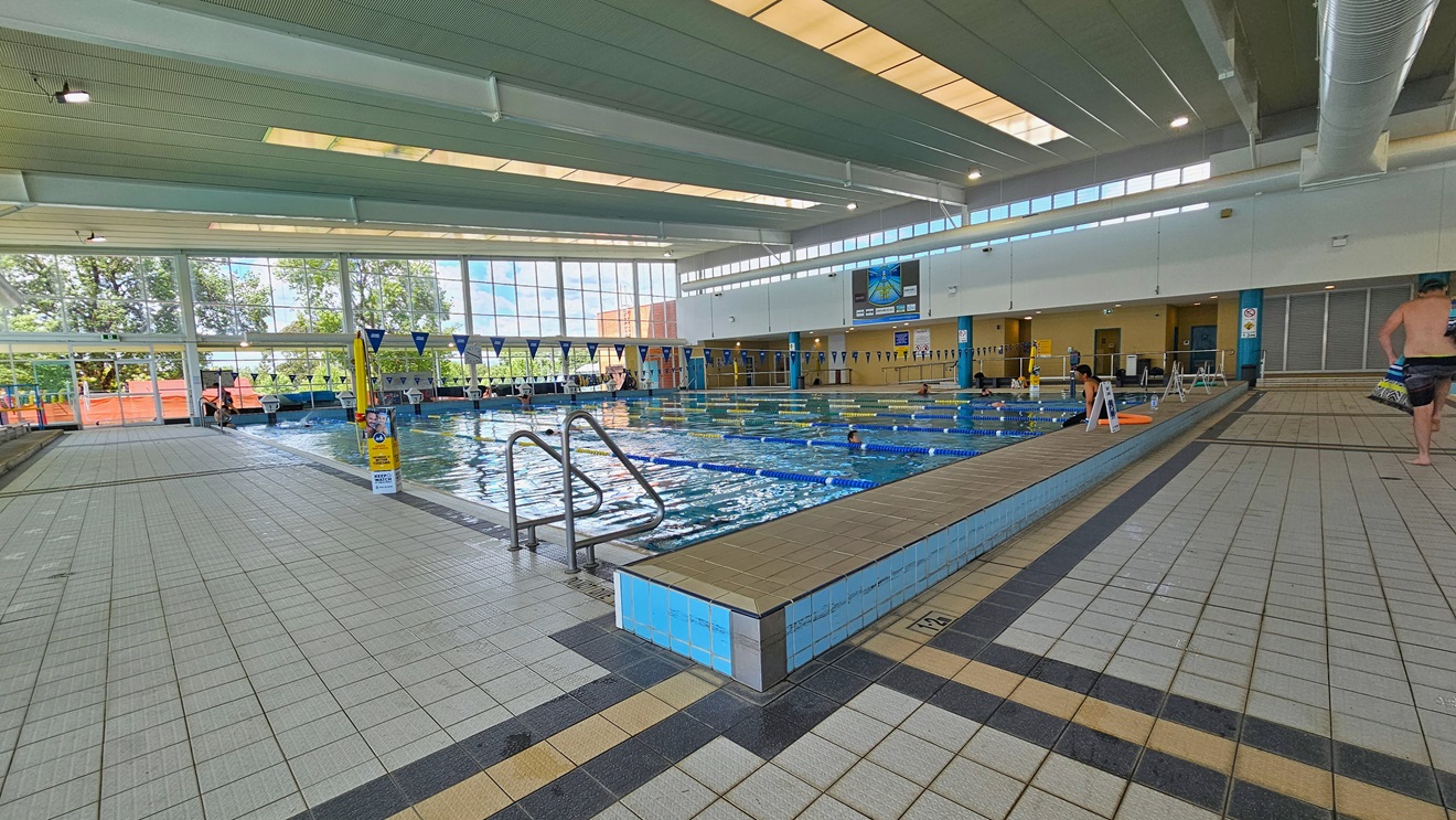 The pool is equipped with handrails, and banners are visible on the pool deck. Floating ropes and blue bunting run across the pool. The raised pool deck features free-standing banners. The left and right pool decks have grey tiles. The surrounding upper wall is made of glass, providing a view of the outside. The high ceiling incorporates strips of transparent roofing, allowing natural sunlight, and large aircon vents. The pool is equipped with handrails, and banners are visible on the pool deck. Floating ropes and blue bunting run across the pool. The raised pool deck features free-standing banners. The left and right pool decks have grey tiles. The surrounding upper wall is made of glass, providing a view of the outside. The high ceiling incorporates strips of transparent roofing, allowing natural sunlight, and large aircon vents.
