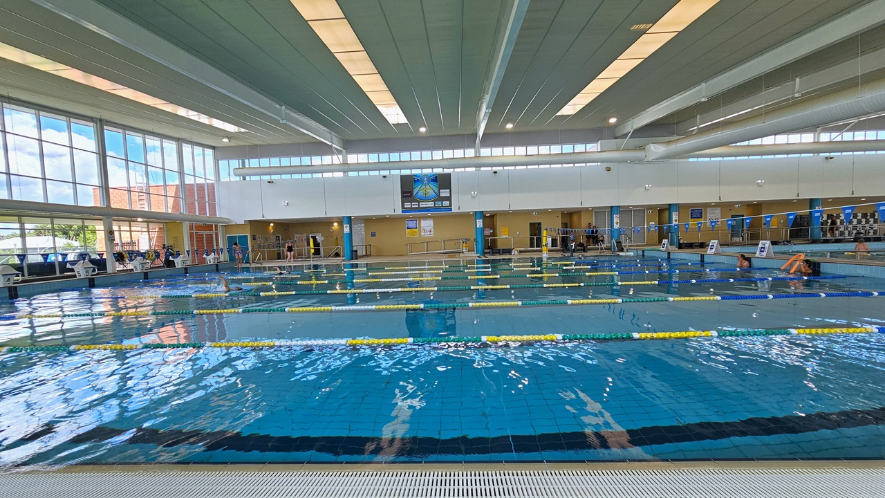 The pool is equipped with colorful floating ropes, and blue buntings run across it. The pool edge features an overflow grid. The raised pool deck has free-standing banners and diving boards. The surrounding upper wall is made of glass, providing a view of the outside with a large poster. The high ceiling incorporates strips of transparent roofing, allowing natural sunlight, and large aircon vents. The pool is equipped with colorful floating ropes, and blue buntings run across it. The pool edge features an overflow grid. The raised pool deck has free-standing banners and diving boards. The surrounding upper wall is made of glass, providing a view of the outside with a large poster. The high ceiling incorporates strips of transparent roofing, allowing natural sunlight, and large aircon vents.