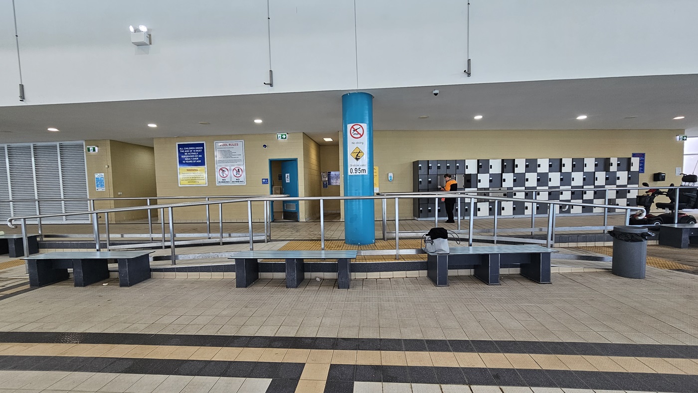 Bench chairs are placed near the ramp entrance, accompanied by rubbish bins. Behind, the locker area in alternating shades of black and white is located on the right. To the left are the accessible bathrooms, with safety posters and stickers affixed to the beige wall. The floor is adorned with cream tiles featuring a black and beige accent. Bench chairs are placed near the ramp entrance, accompanied by rubbish bins. Behind, the locker area in alternating shades of black and white is located on the right. To the left are the accessible bathrooms, with safety posters and stickers affixed to the beige wall. The floor is adorned with cream tiles featuring a black and beige accent.