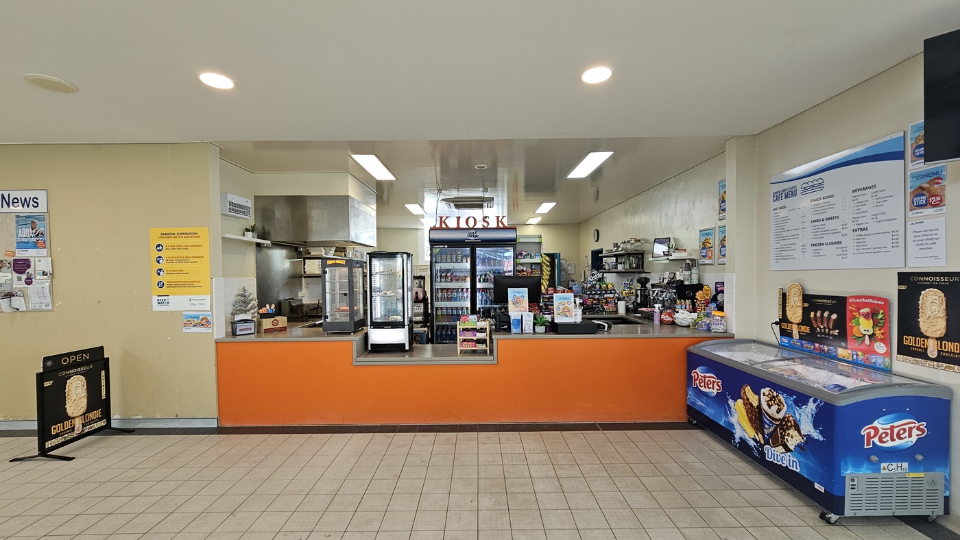 The kiosk showcases a low orange reception counter with food racks and items. Behind it, a fridge stores cold drinks, and display racks hold snacks. To the right, an ice cream freezer is positioned. Advert posters and facility reminders are affixed to the walls, with an advert poster standing on the left. The kiosk showcases a low orange reception counter with food racks and items. Behind it, a fridge stores cold drinks, and display racks hold snacks. To the right, an ice cream freezer is positioned. Advert posters and facility reminders are affixed to the walls, with an advert poster standing on the left.
