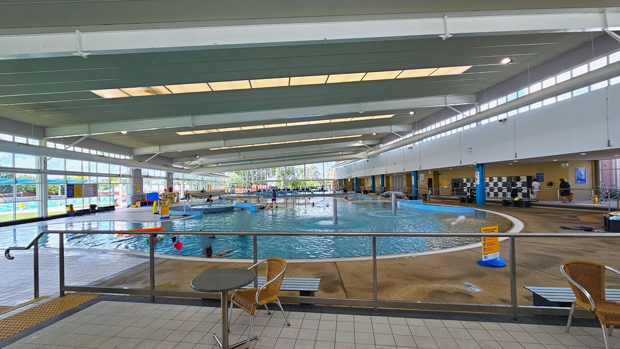 Next to the Leisure Pool is an elevated area near the cafe with movable tables and chairs. On the right, steps with handrails and truncated domes are present, along with bench chairs. The Leisure Pool is in front, and further to the right is the locker area. The upper wall is glass, offering an outside view with a large poster. The high ceiling includes strips of transparent roofing for natural sunlight and large aircon vents. Next to the Leisure Pool is an elevated area near the cafe with movable tables and chairs. On the right, steps with handrails and truncated domes are present, along with bench chairs. The Leisure Pool is in front, and further to the right is the locker area. The upper wall is glass, offering an outside view with a large poster. The high ceiling includes strips of transparent roofing for natural sunlight and large aircon vents.