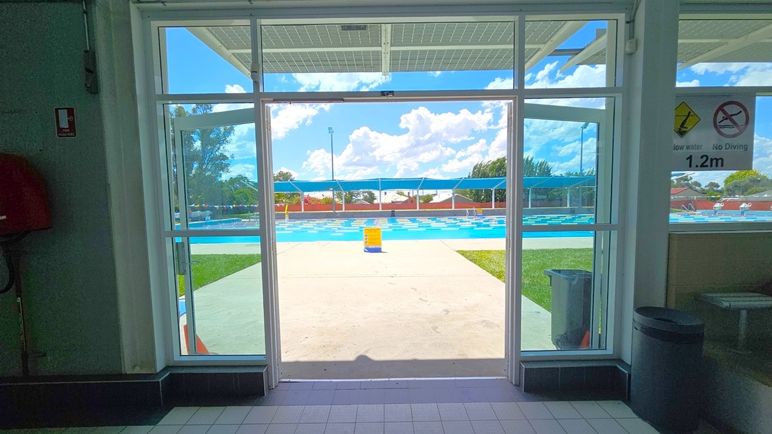 Looking through a glass door, a swimming pool is visible outside. The pool area has a tiled path leading to it, with grass on the right. A person is sitting by the pool, and there are blue shade structures above. Signs on the door indicate "1.2m" depth and "No Diving". Inside, the floor is tiled, and a rubbish bin is on the right. The ceiling inside is not visible. Looking through a glass door, a swimming pool is visible outside. The pool area has a tiled path leading to it, with grass on the right. A person is sitting by the pool, and there are blue shade structures above. Signs on the door indicate "1.2m" depth and "No Diving". Inside, the floor is tiled, and a rubbish bin is on the right. The ceiling inside is not visible.