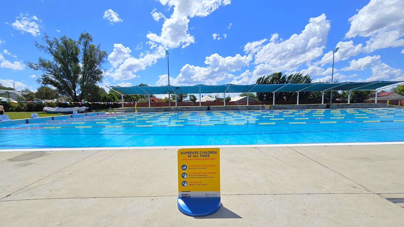 A large outdoor swimming pool with clear blue water is in the centre. In the foreground, a yellow sign on the pool deck advises supervising children. The pool is divided into lanes with floating markers. In the background, there are blue shade structures and a few trees. The ground is paved with light-coloured concrete. A large outdoor swimming pool with clear blue water is in the centre. In the foreground, a yellow sign on the pool deck advises supervising children. The pool is divided into lanes with floating markers. In the background, there are blue shade structures and a few trees. The ground is paved with light-coloured concrete.