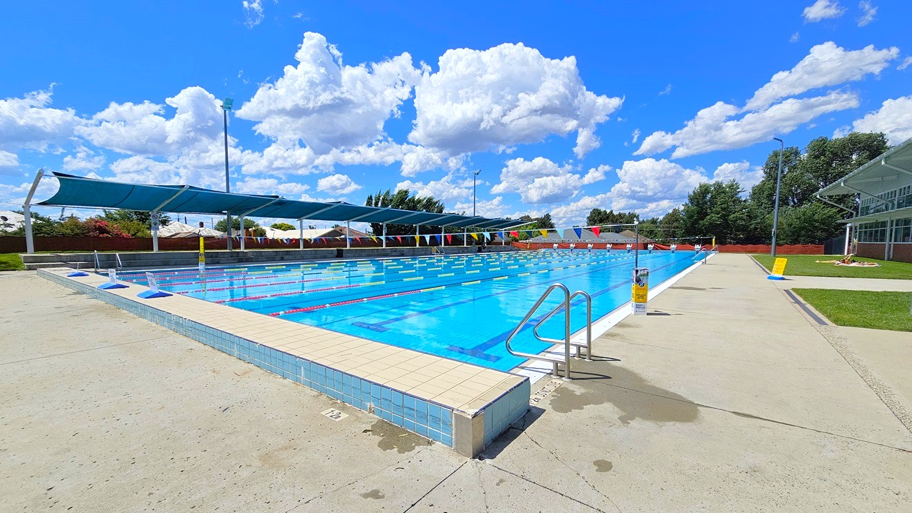 Outdoor swimming pool with clear blue water, surrounded by a concrete deck. On the left, there are several shaded areas with blue canopies. A metal handrail descends into the pool. On the right, a grassy area and a building with large windows are visible. The pool is divided into lanes with red and white lane markers. In the background, trees and a fence line the perimeter. The sky is filled with fluffy white clouds. Outdoor swimming pool with clear blue water, surrounded by a concrete deck. On the left, there are several shaded areas with blue canopies. A metal handrail descends into the pool. On the right, a grassy area and a building with large windows are visible. The pool is divided into lanes with red and white lane markers. In the background, trees and a fence line the perimeter. The sky is filled with fluffy white clouds.