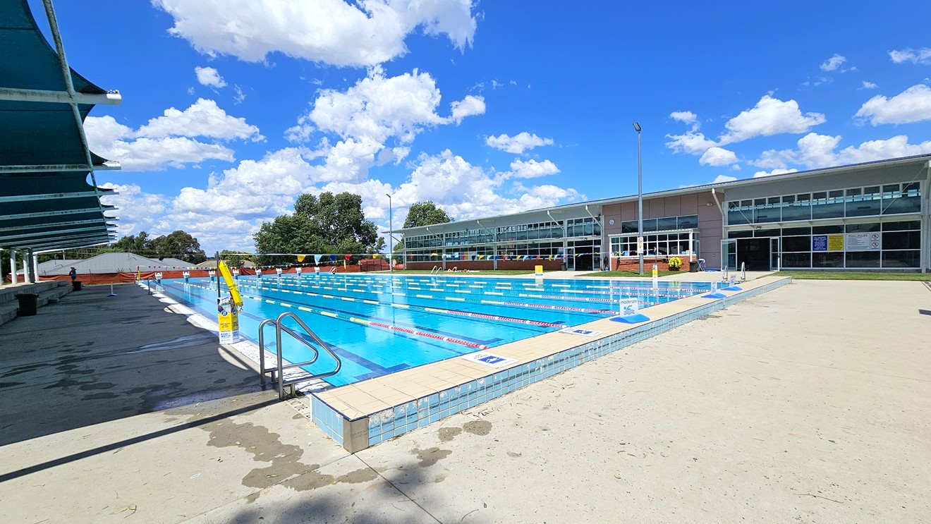 An outdoor swimming pool with clear blue water and lane dividers. On the left, there are shaded seating areas with dark blue canopies. The right side features a modern building with large glass windows. In the background, trees and a few clouds are visible. The pool deck is light-coloured concrete with a metal handrail leading into the water. An outdoor swimming pool with clear blue water and lane dividers. On the left, there are shaded seating areas with dark blue canopies. The right side features a modern building with large glass windows. In the background, trees and a few clouds are visible. The pool deck is light-coloured concrete with a metal handrail leading into the water.