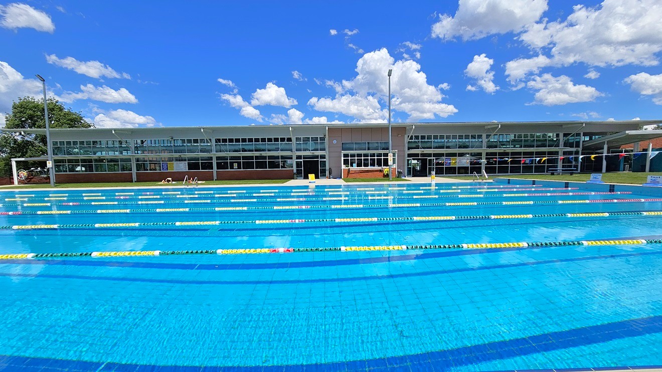 An outdoor swimming pool with multiple lanes marked by yellow and black lane dividers. In the background, a modern building with large glass windows stretches across the image. A few people are visible near the building, possibly sunbathing or resting. The pool area is surrounded by a grassy lawn, and there are tall lights on poles. The water in the pool is clear and blue, reflecting the surroundings. An outdoor swimming pool with multiple lanes marked by yellow and black lane dividers. In the background, a modern building with large glass windows stretches across the image. A few people are visible near the building, possibly sunbathing or resting. The pool area is surrounded by a grassy lawn, and there are tall lights on poles. The water in the pool is clear and blue, reflecting the surroundings.