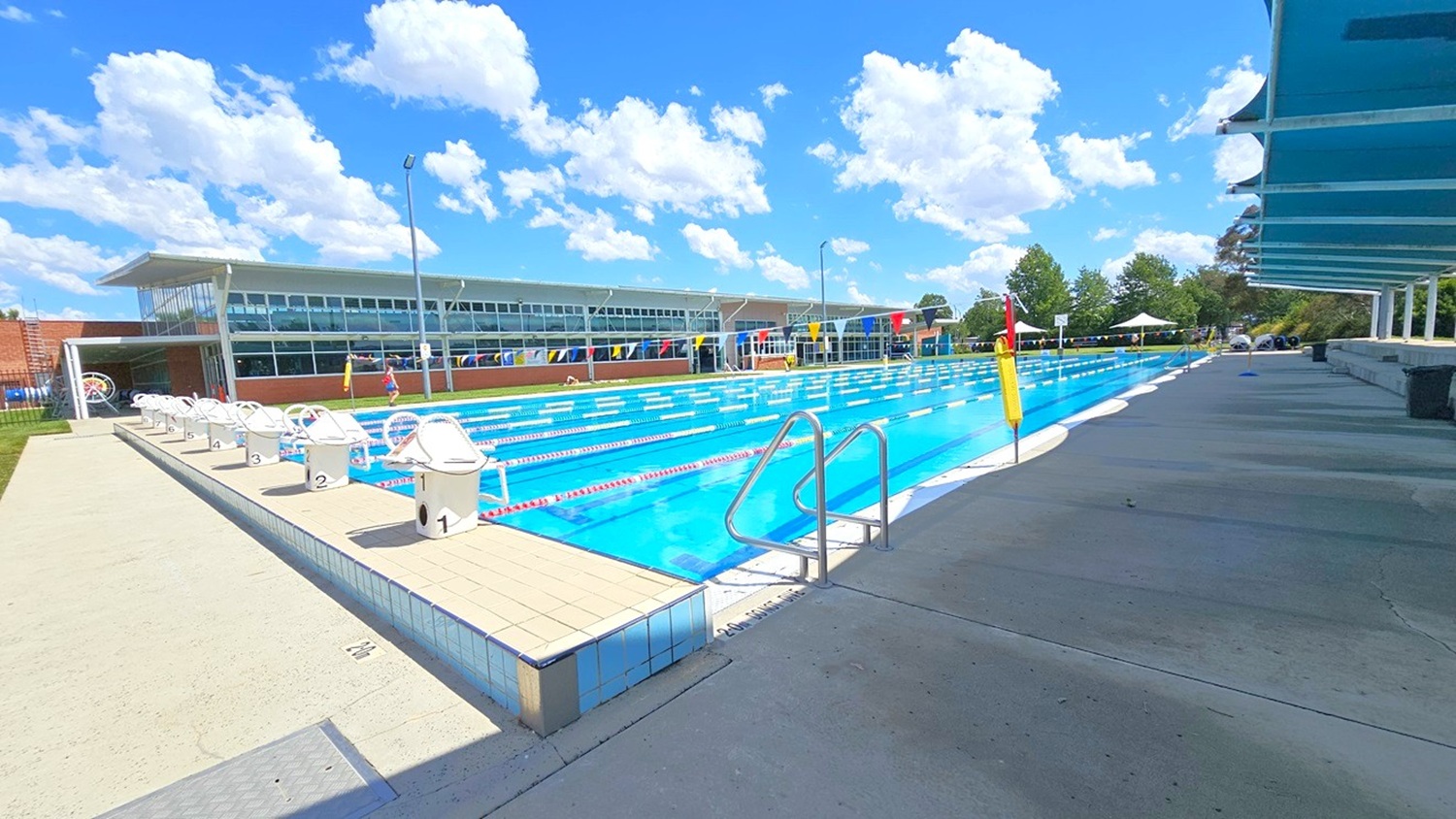 An outdoor swimming pool with multiple lanes, surrounded by a concrete deck. On the left, there are starting blocks numbered sequentially. A modern building with large windows is in the background. On the right, a covered walkway provides shade. Trees and a small tent are visible in the distance. The pool is equipped with lane dividers and a metal ladder for entry. An outdoor swimming pool with multiple lanes, surrounded by a concrete deck. On the left, there are starting blocks numbered sequentially. A modern building with large windows is in the background. On the right, a covered walkway provides shade. Trees and a small tent are visible in the distance. The pool is equipped with lane dividers and a metal ladder for entry.