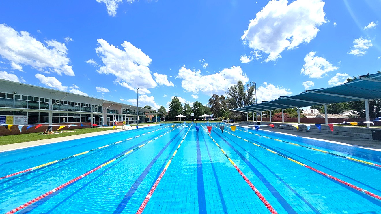An outdoor swimming pool with multiple lanes, each marked by red and yellow lane dividers. On the left, a modern building with large windows and colourful wall accents. On the right, a series of blue shade structures providing cover. In the background, trees and white umbrellas are visible. The pool is surrounded by a paved area and grass. An outdoor swimming pool with multiple lanes, each marked by red and yellow lane dividers. On the left, a modern building with large windows and colourful wall accents. On the right, a series of blue shade structures providing cover. In the background, trees and white umbrellas are visible. The pool is surrounded by a paved area and grass.