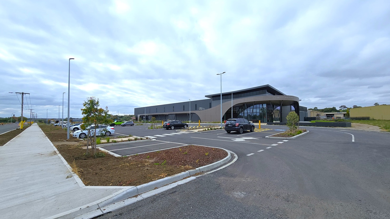 A car park with parked vehicles. Concrete pavement separates it from the road. Accessible parking is marked with signs and yellow bollards. A concrete plant box has plants and small trees. Next to it is a modern facility building, with a grass area to the right. A car park with parked vehicles. Concrete pavement separates it from the road. Accessible parking is marked with signs and yellow bollards. A concrete plant box has plants and small trees. Next to it is a modern facility building, with a grass area to the right.