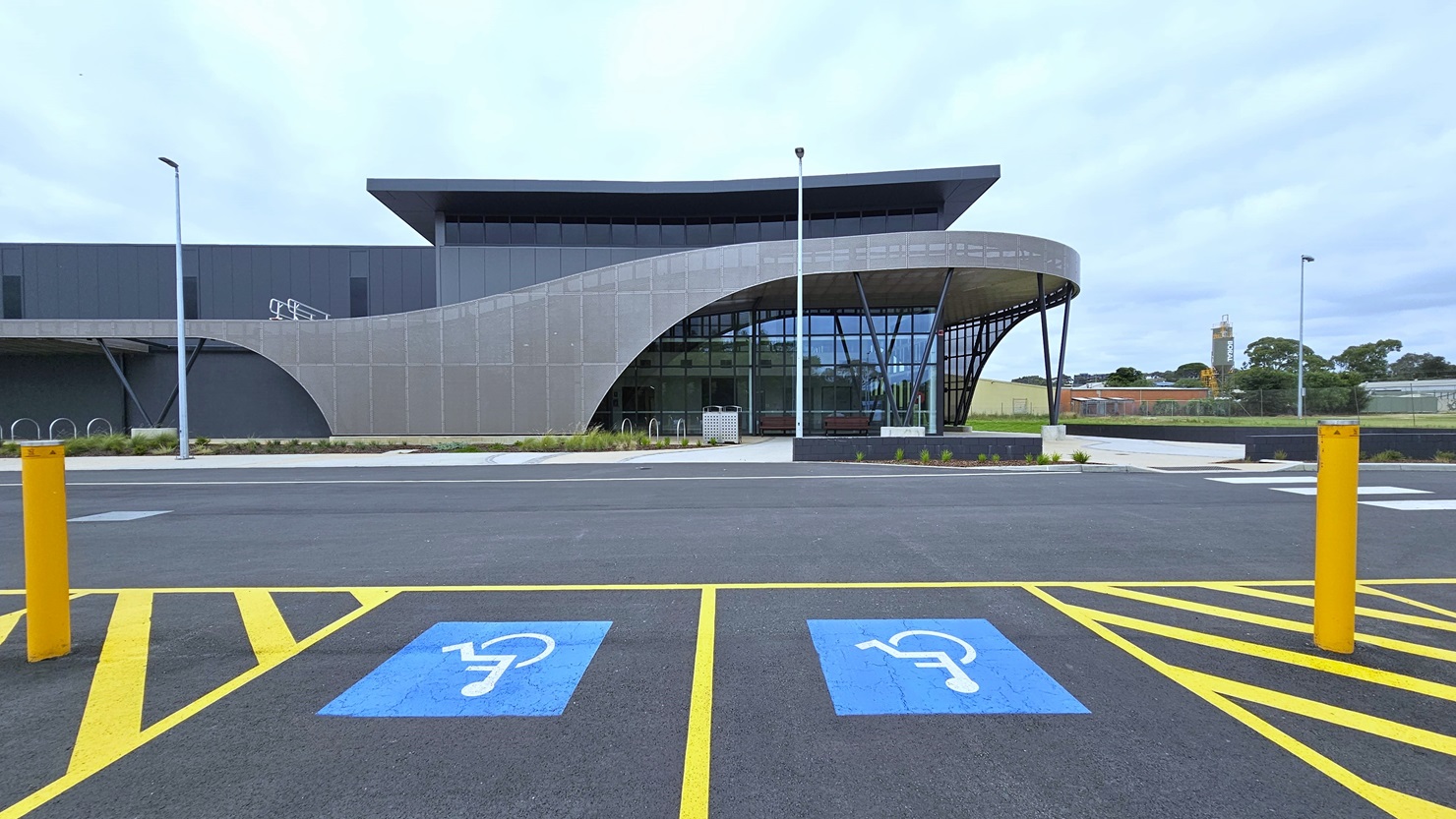 Accessible car parking spaces are marked with signs and yellow bollards. Adjacent is a modern facility building made of concrete, glass, and beams. A grass area lies to the left, bordered by tall lamp posts along the road. Bike racks are positioned at the sides of the building. A concrete plant box hosts plants and small trees. Accessible car parking spaces are marked with signs and yellow bollards. Adjacent is a modern facility building made of concrete, glass, and beams. A grass area lies to the left, bordered by tall lamp posts along the road. Bike racks are positioned at the sides of the building. A concrete plant box hosts plants and small trees.